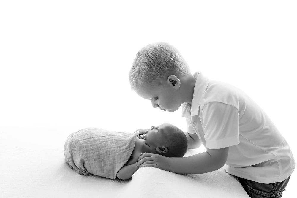 Black and white photo of young boy tenderly kissing sleeping newborn sibling wrapped in swaddle.