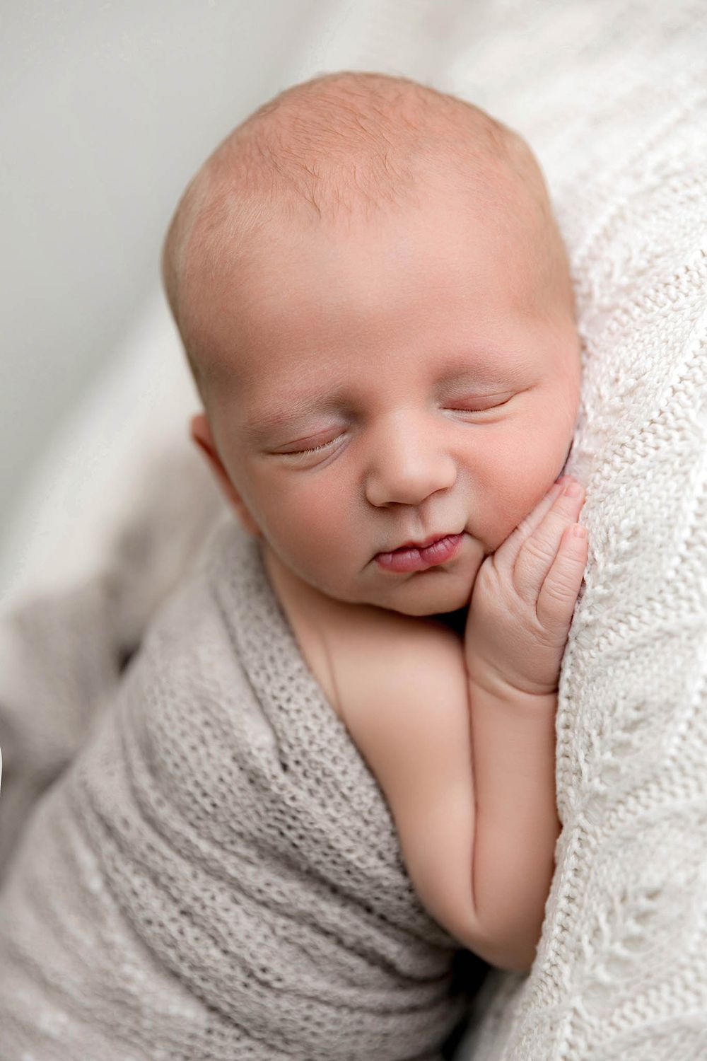 Sleeping newborn baby wrapped in soft gray knit blanket with hands near face.