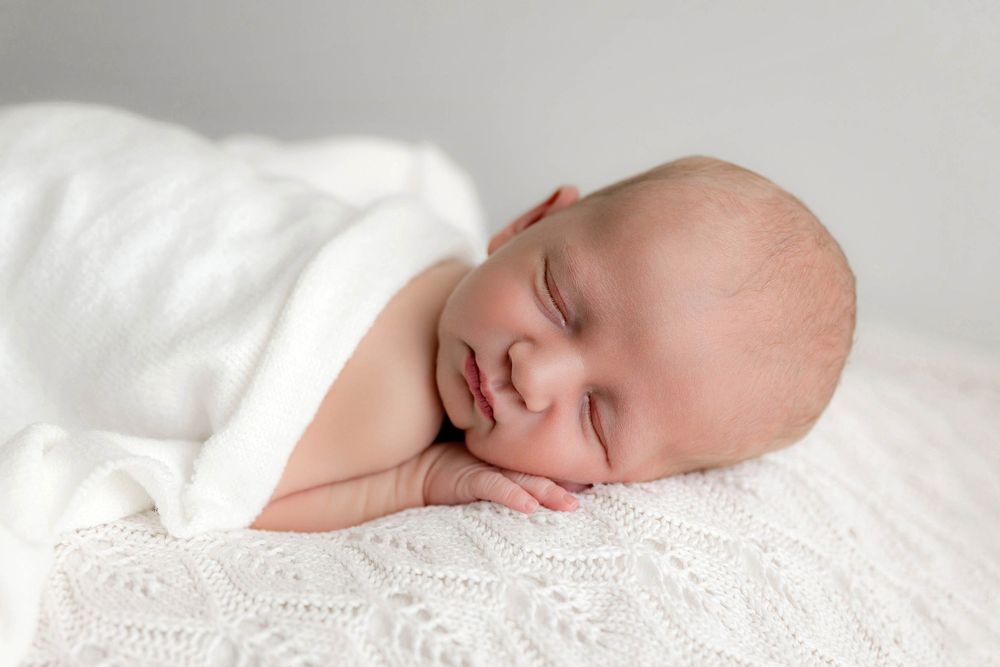Peaceful newborn baby sleeping wrapped in a soft white blanket on a white knit surface.