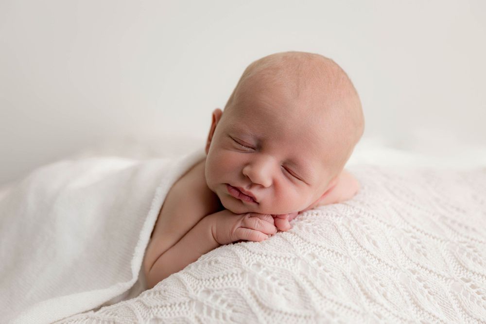Peaceful newborn baby sleeping on white knit blanket with hands tucked under chin in soft natural light.