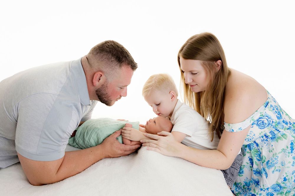 A family with a toddler and swaddled newborn poses together on a white surface in a bright studio photo.