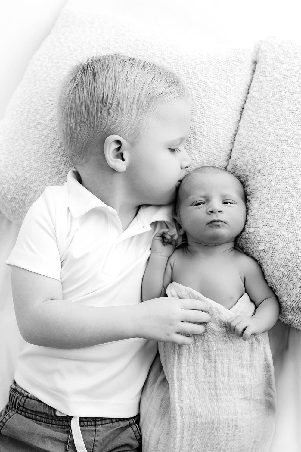 Black and white photo of a young boy kissing his newborn sibling wrapped in a soft blanket on a couch.