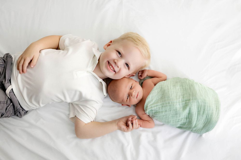 Smiling toddler lying on white bed with sleeping newborn baby wrapped in green swaddle blanket.