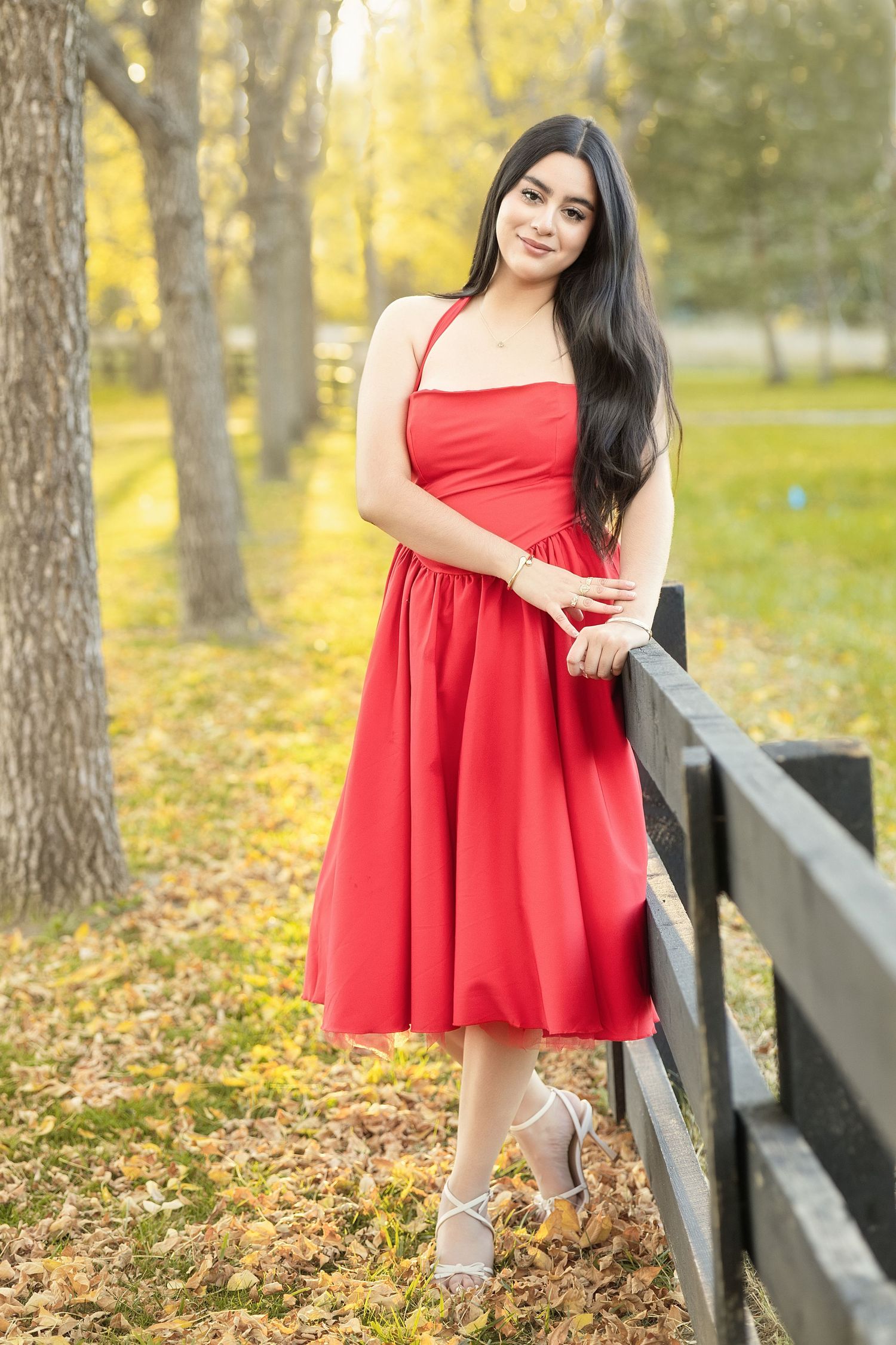 Young woman in red midi dress leans on wooden fence in autumn park with golden leaves and trees.