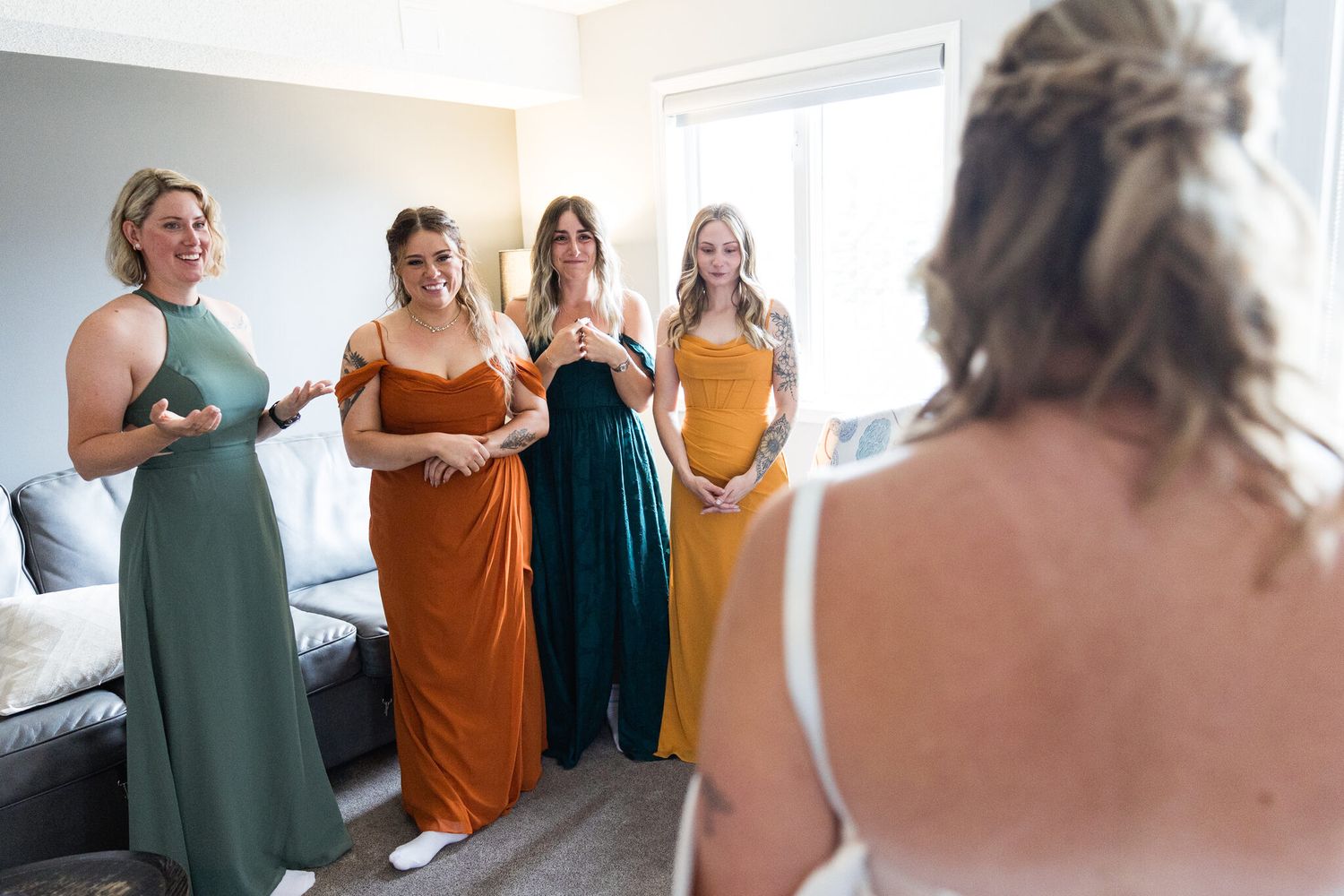 Bridesmaids in sage, rust and mustard dresses react to the bride before her Canmore wedding