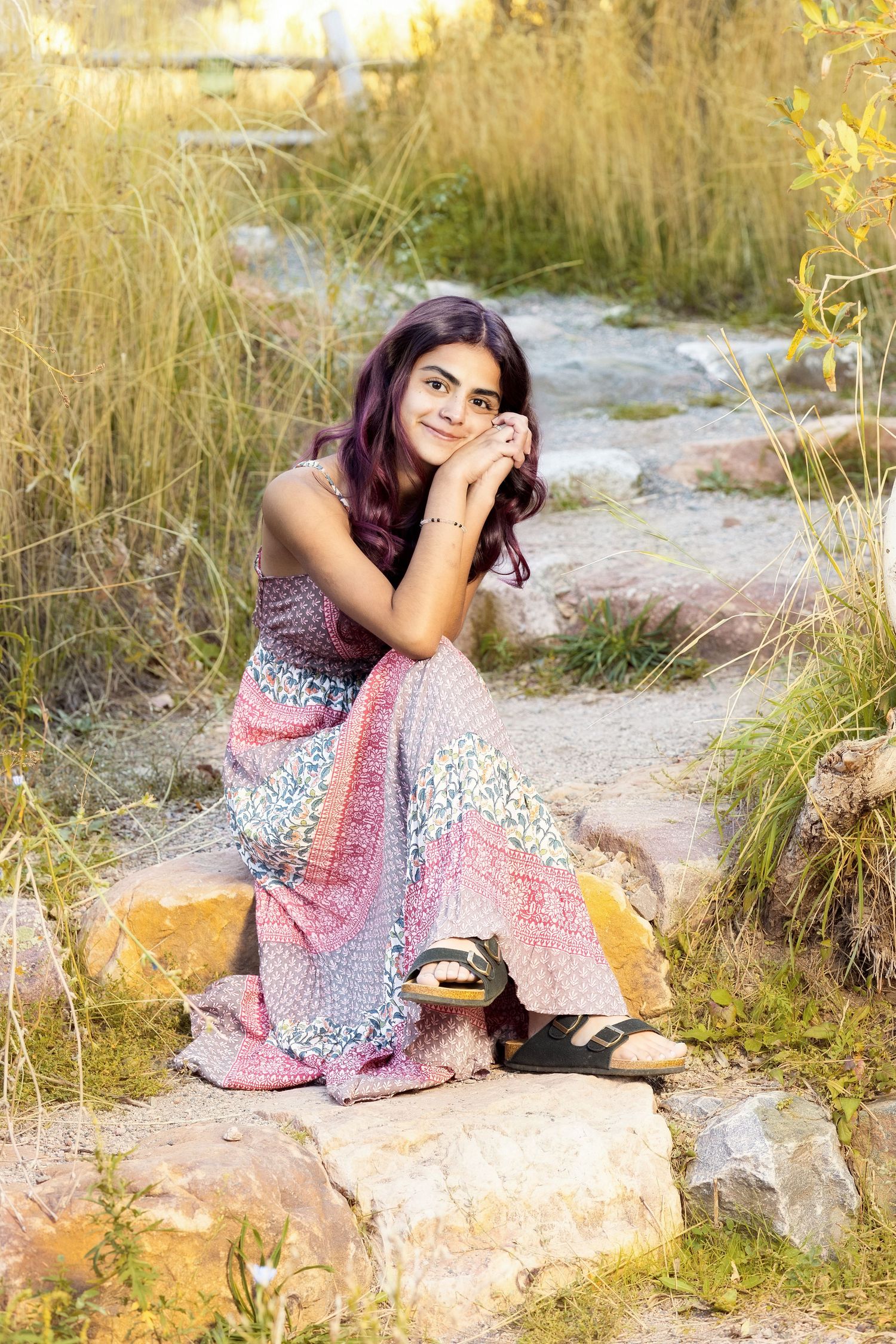 A young woman with purple hair sits on rocks in a boho floral maxi dress and sandals, surrounded by tall golden grass.