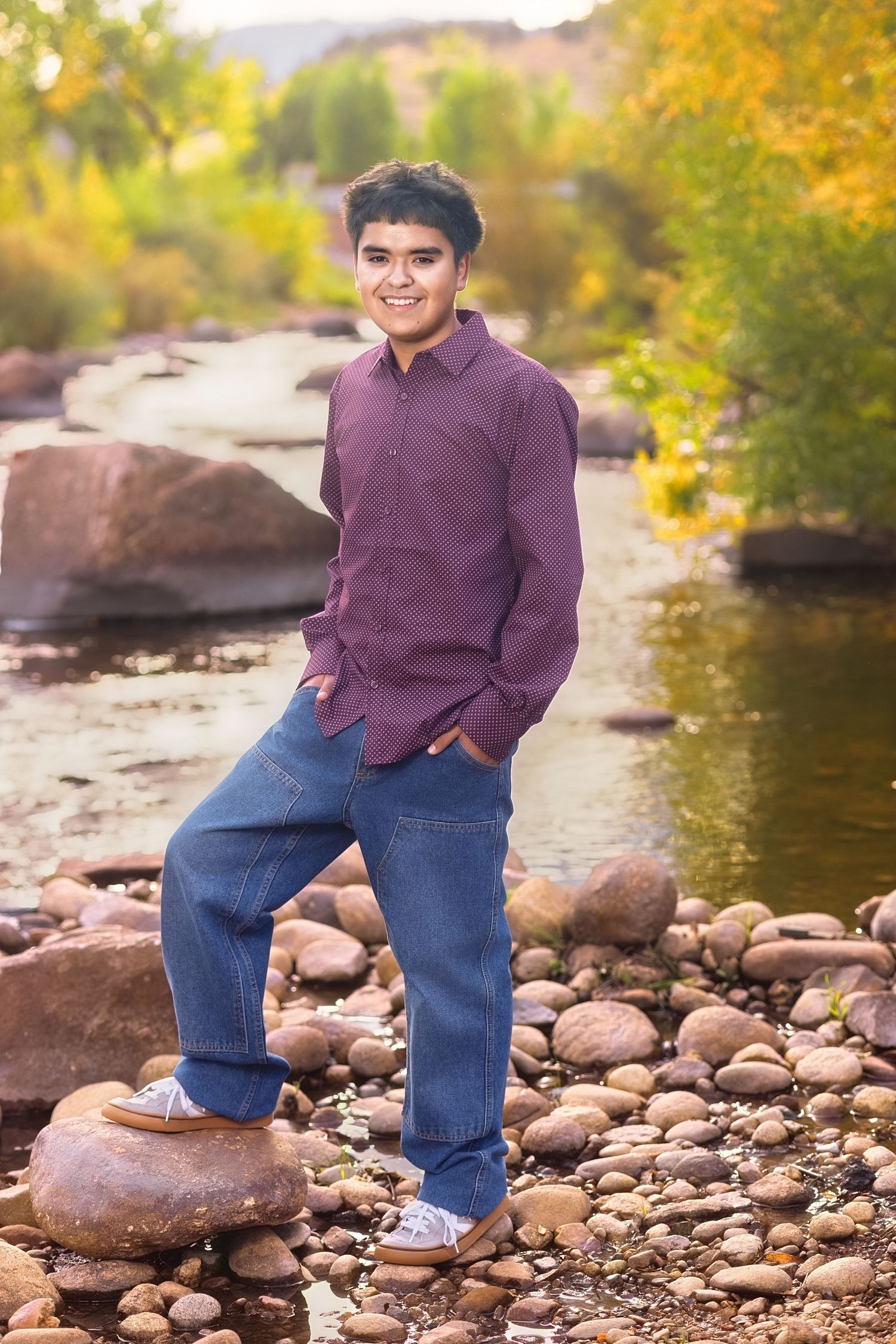 Smiling teen boy in purple shirt and jeans poses on river rocks with autumn trees in background.