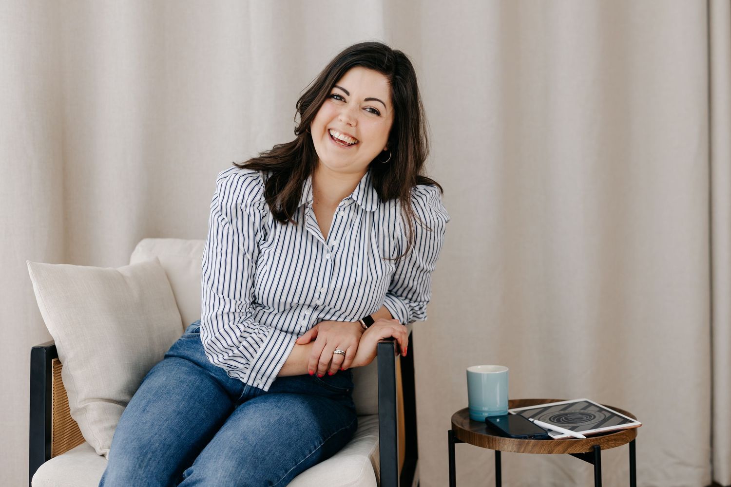 Smiling woman in striped shirt sitting relaxed in a chair next to a small side table with a mug.