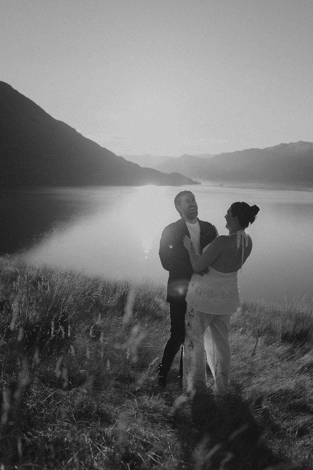 Black and white photo of a couple embracing at sunset by a serene mountain lake, dressed in wedding attire.