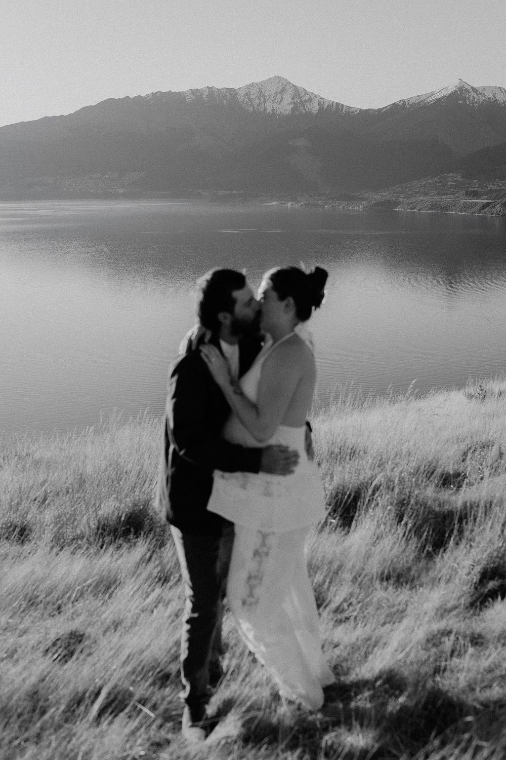 Black and white photo of a couple kissing on a hillside with a lake and snow-capped mountains in the background.