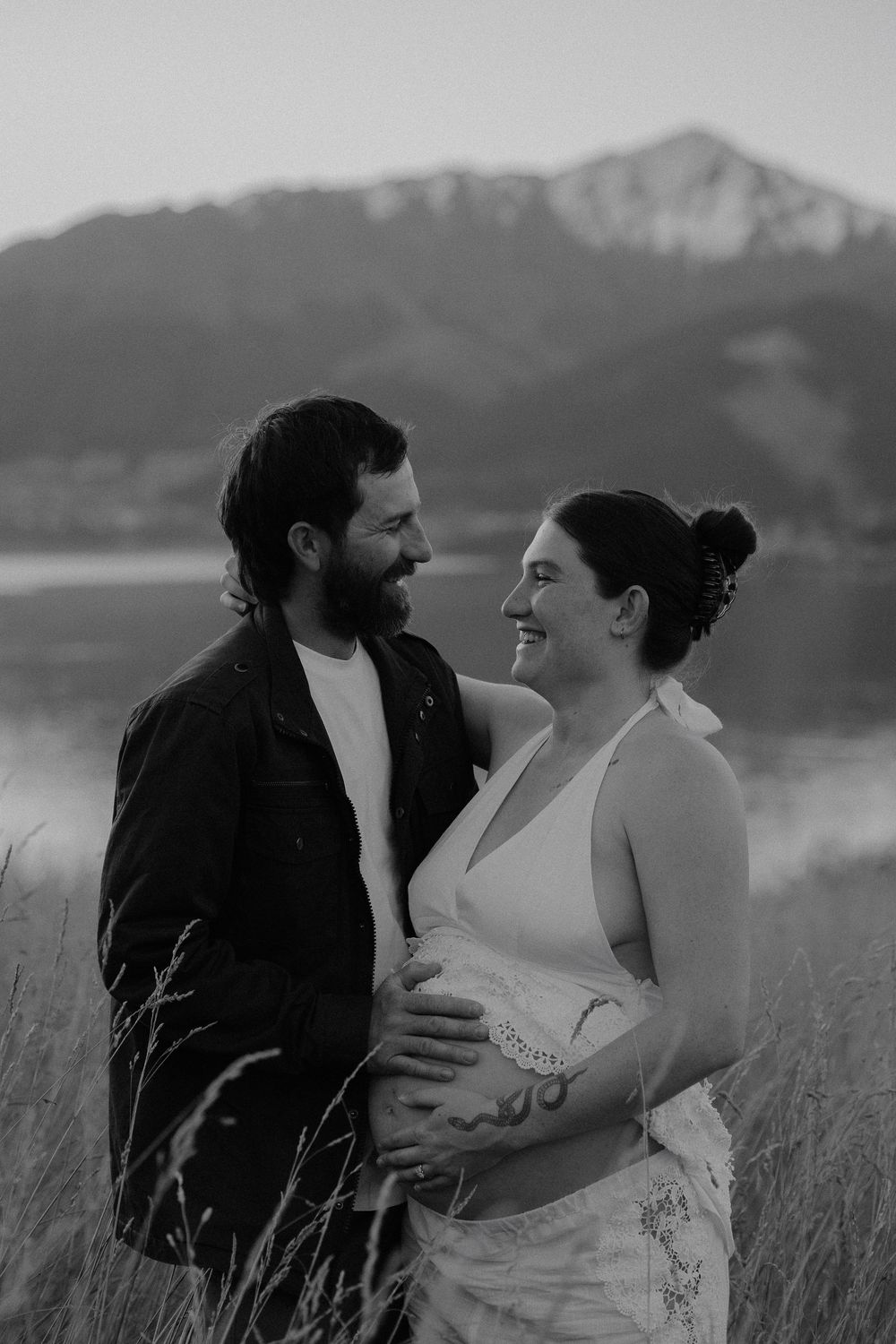 Black and white photo of a smiling pregnant couple embracing in a field with a mountain lake backdrop.