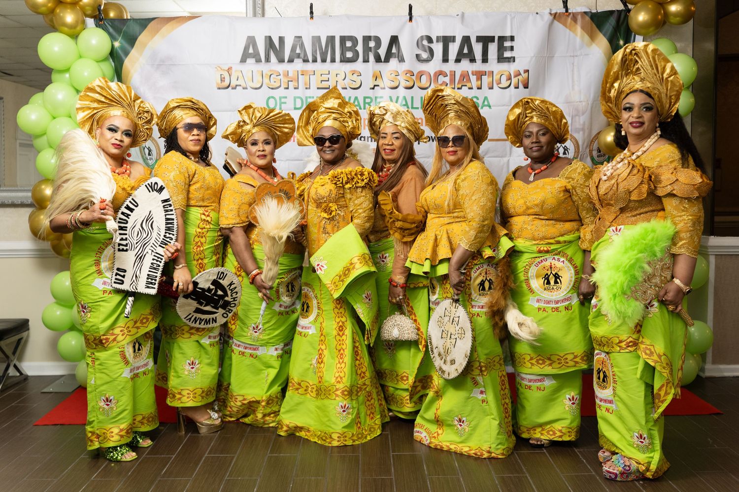 Group of women in matching gold and green traditional attire with headpieces posing before an Anambra State Daughters Association banner.