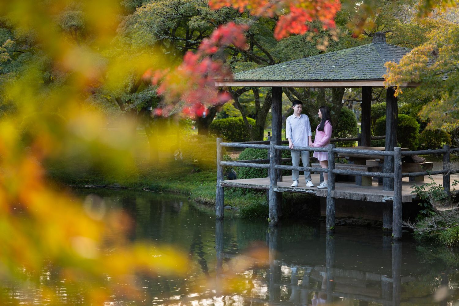 Couple stands on a wooden pavilion deck beside a calm pond, surrounded by colorful red and orange autumn foliage in a peaceful garden.