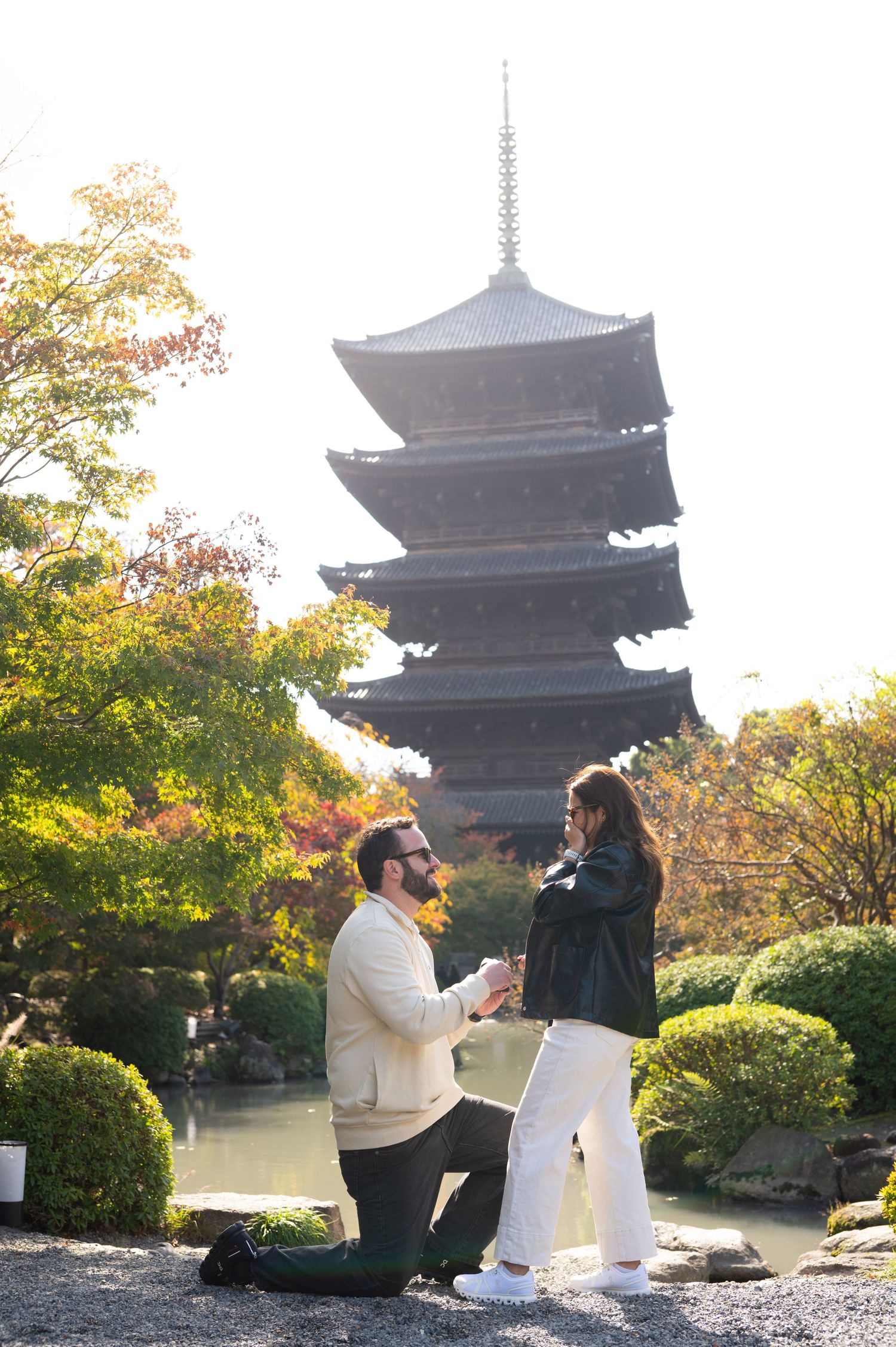 Man kneeling on one knee proposing to a woman beside a pond with a five-story Japanese pagoda and autumn foliage behind them.