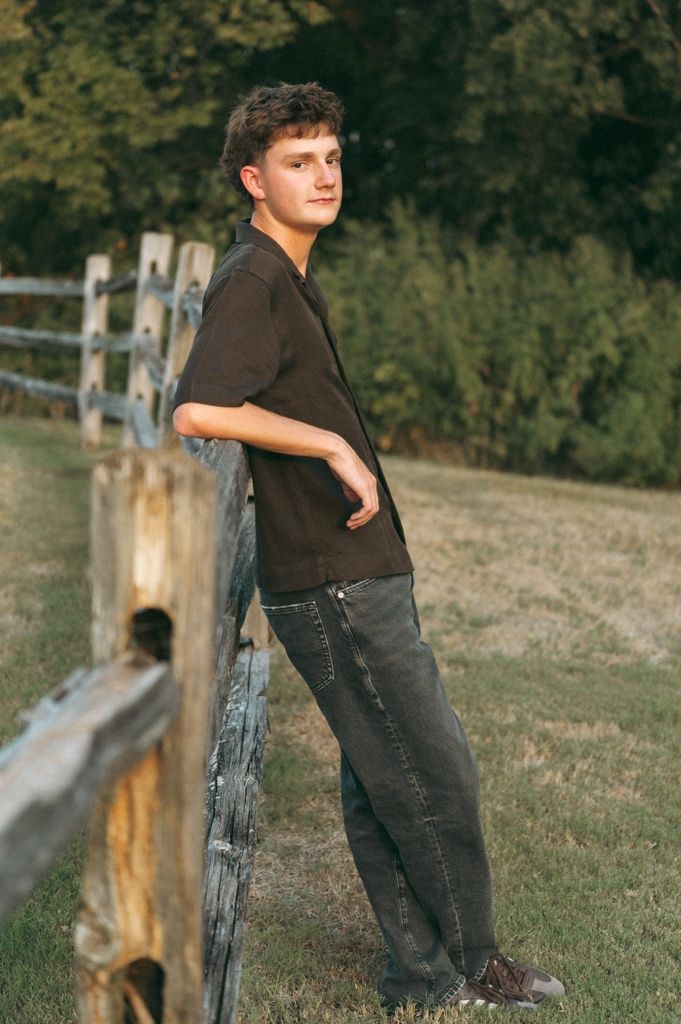 Young man in dark shirt and jeans leaning against rustic wooden fence in a sunny field with green trees.