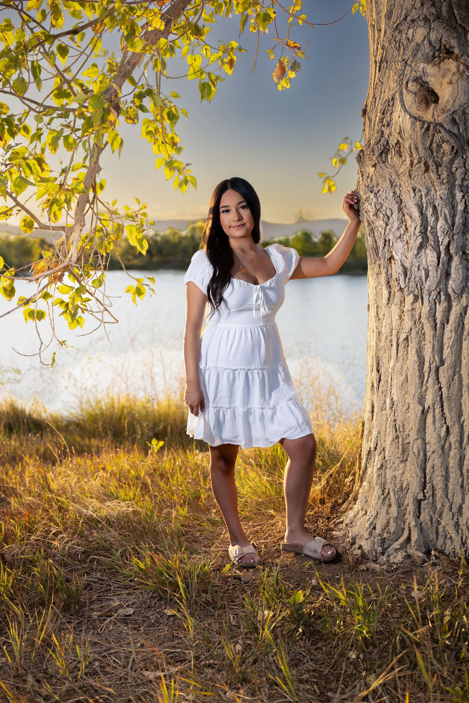 Young woman in white dress leaning against tree by lake at golden hour sunset.