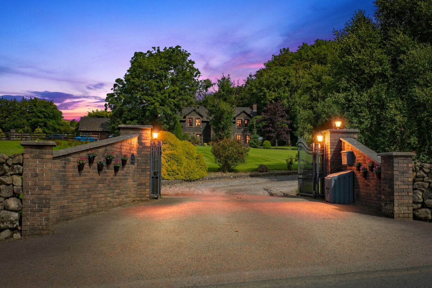 Grand estate entrance at dusk with stone pillars, ornate iron gates, lanterns, and tree-lined driveway.