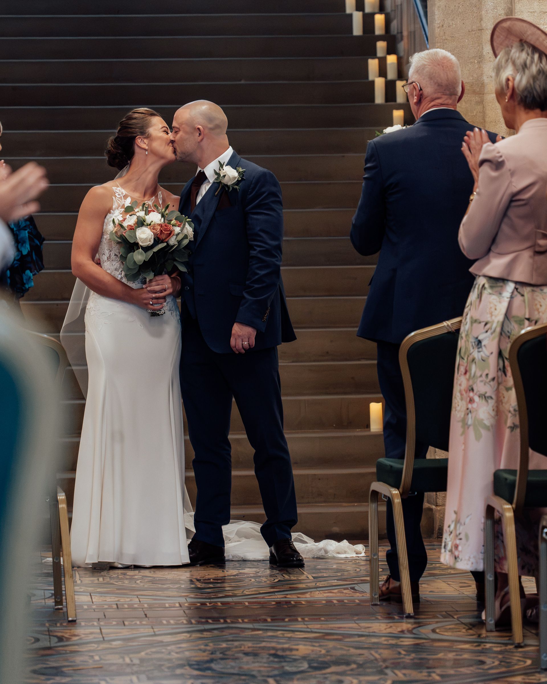 Bride in a white gown holding a bouquet kisses her groom in a navy suit beside a staircase as wedding guests look on.