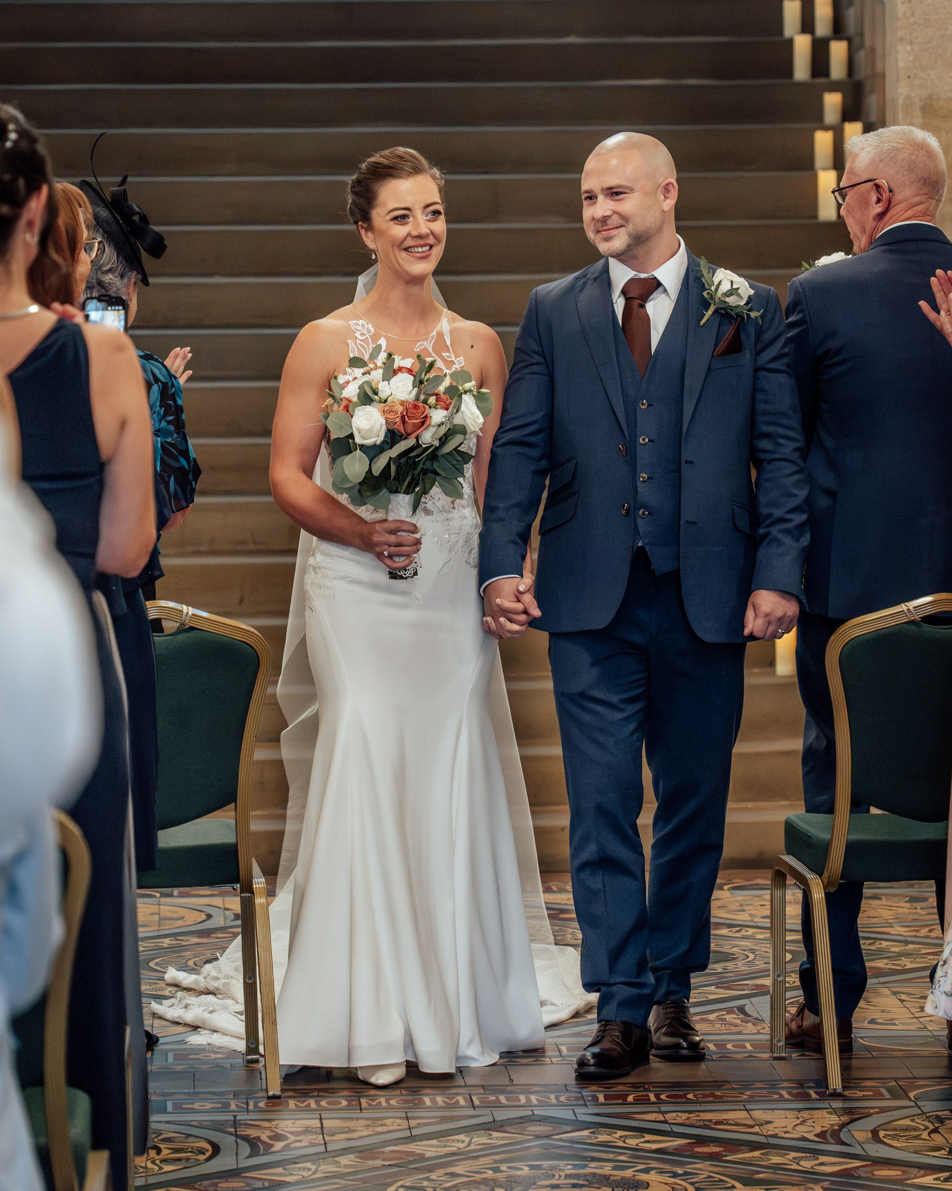 Smiling bride in a white gown holding a bouquet walks hand in hand with the groom in a navy suit down a wedding aisle.