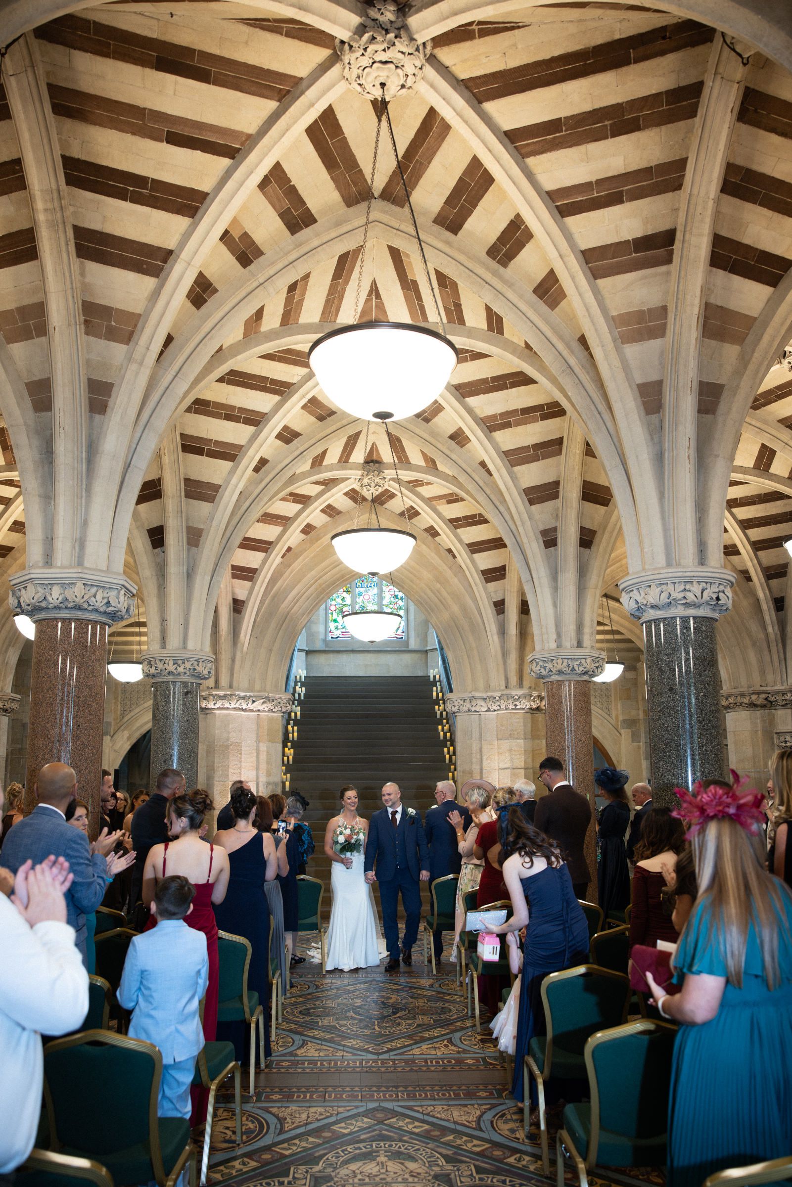 Bride and groom walk down the aisle of an ornate vaulted hall as wedding guests in colorful attire watch from both sides.