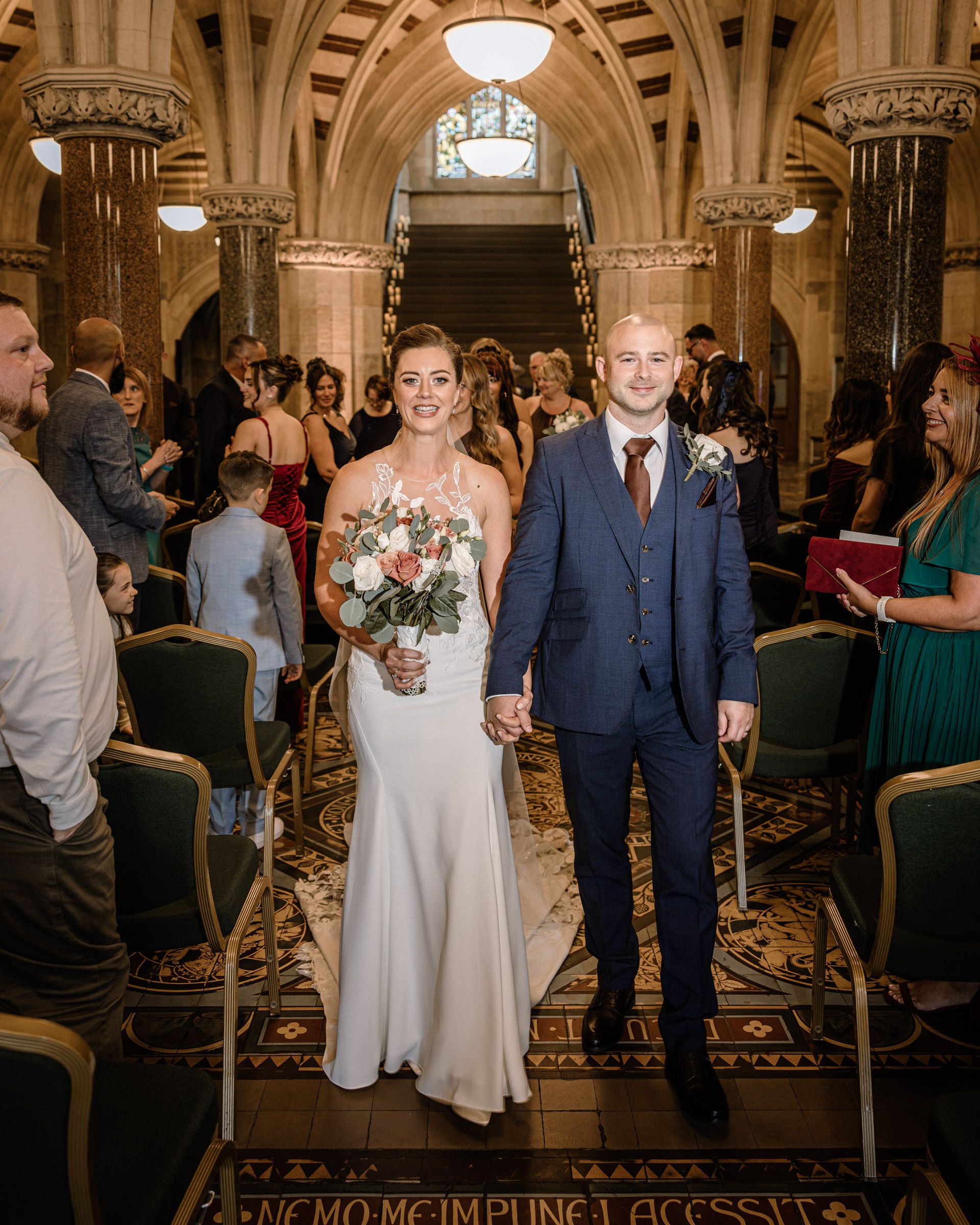 Smiling bride in a white gown holding a bouquet walks hand in hand with the groom in a navy suit down the aisle past wedding guests.