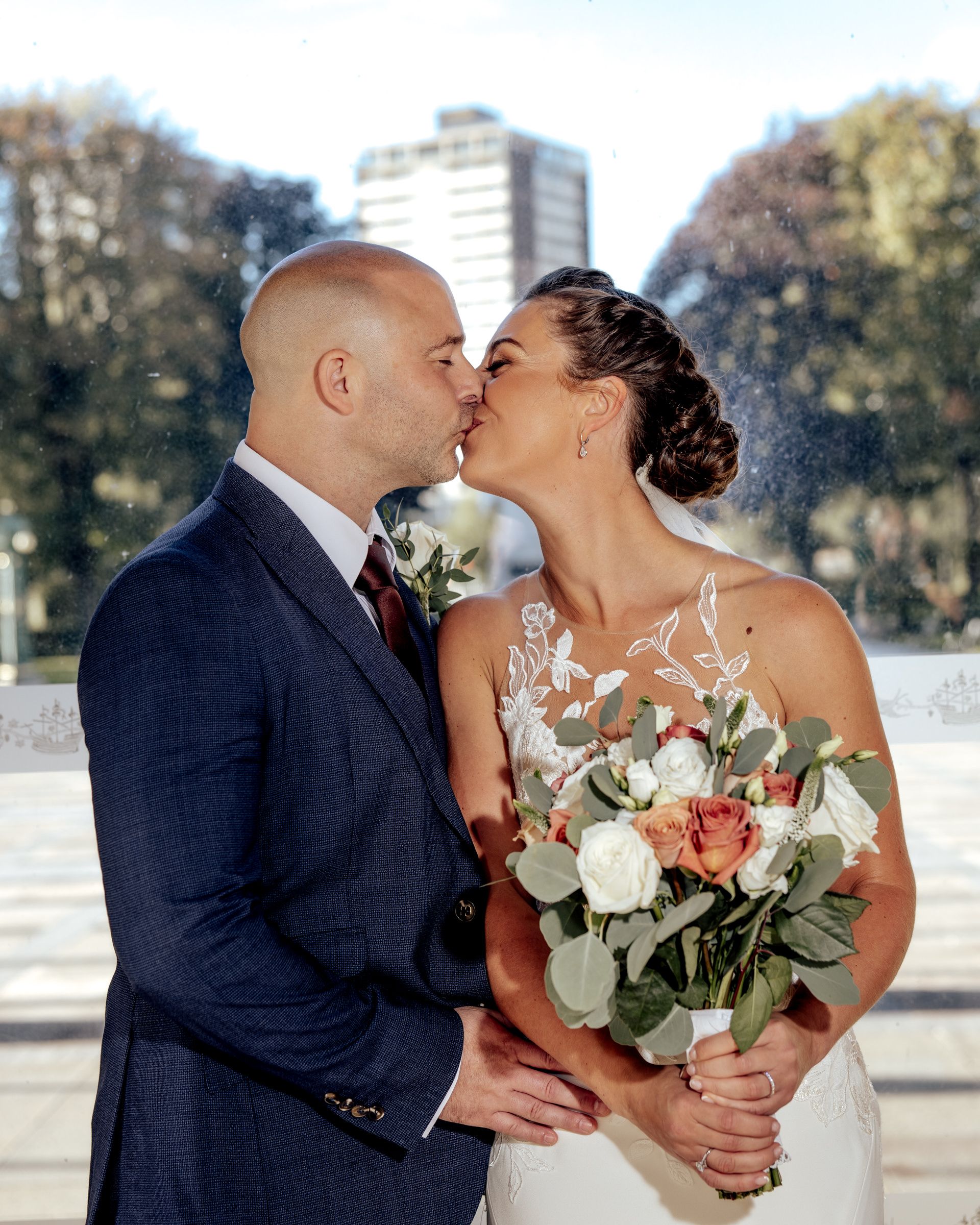 A bride and groom share a kiss outdoors, she holding a bouquet of white and peach roses with greenery, he in a navy suit.