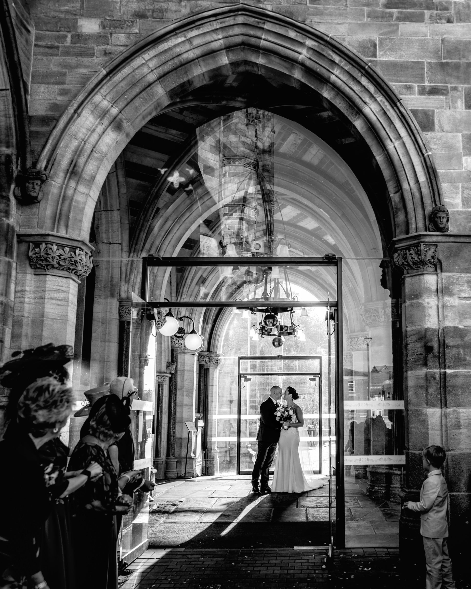 Black and white photo of a bride and groom embracing under a grand Gothic stone archway as wedding guests watch from the side.