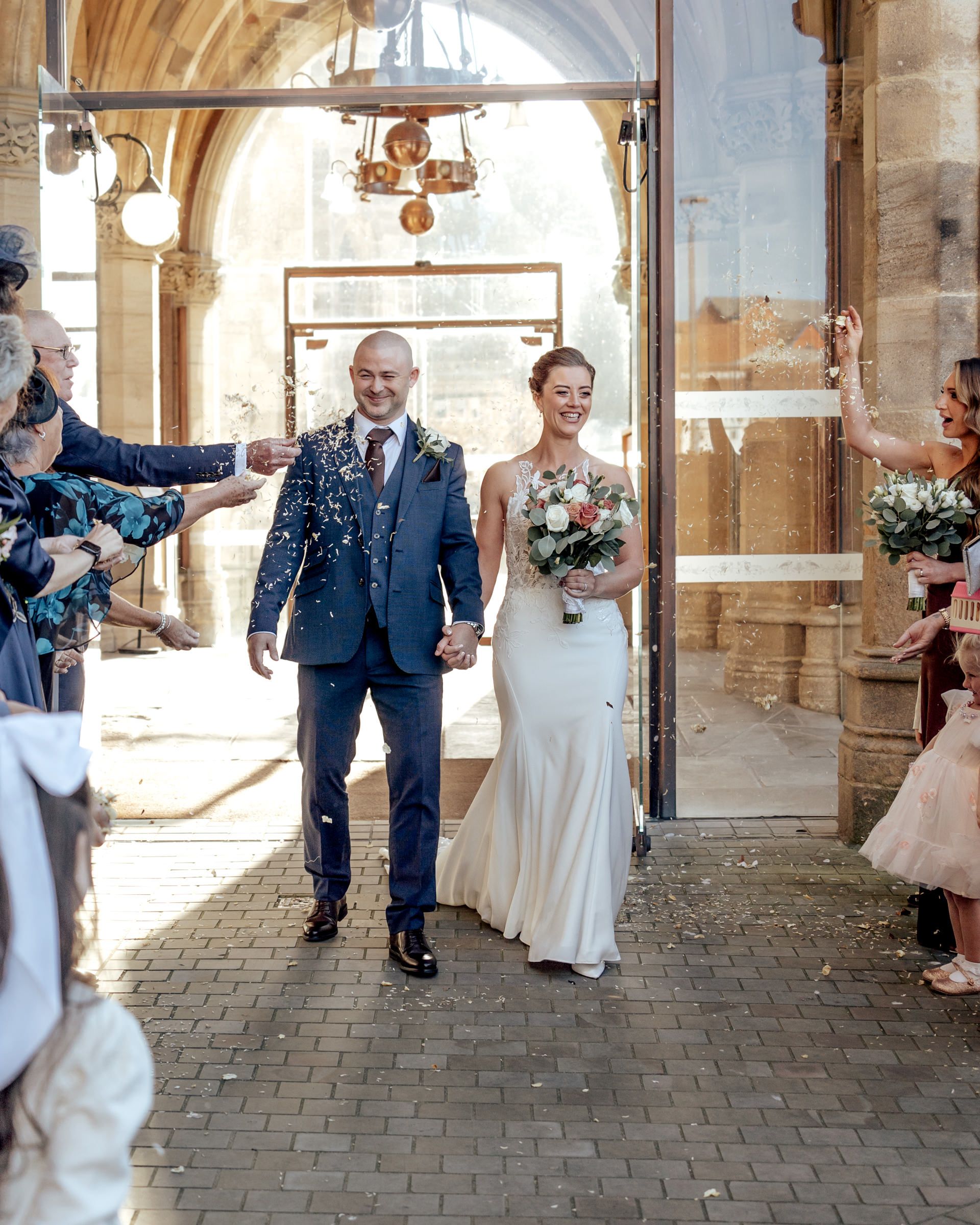 A smiling bride in a white gown and groom in a blue suit walk hand in hand through confetti as wedding guests cheer outside a stone archway.