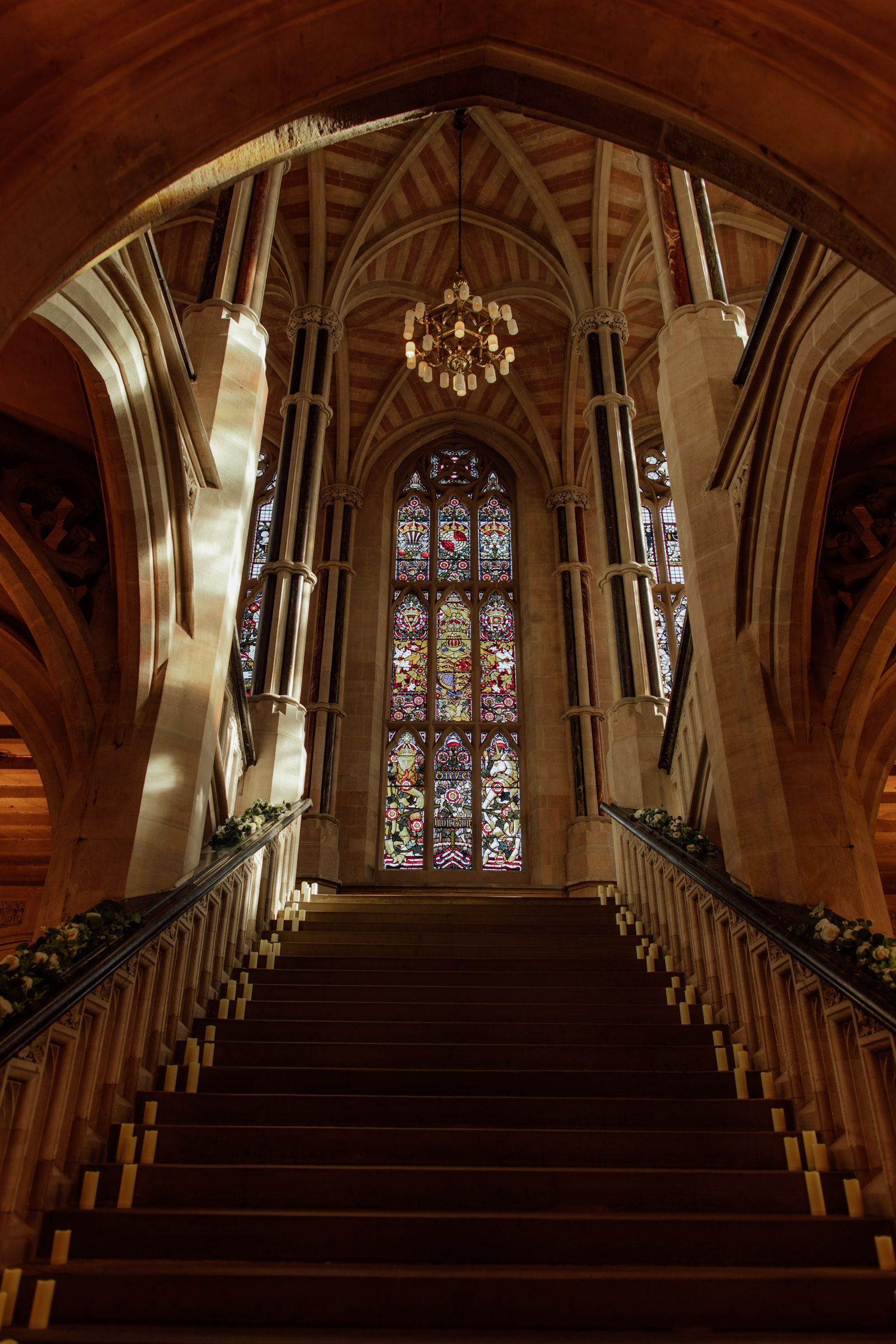 Grand gothic staircase with ornate stained glass window, vaulted ceiling, chandelier and floral garland decorations on the balustrades.