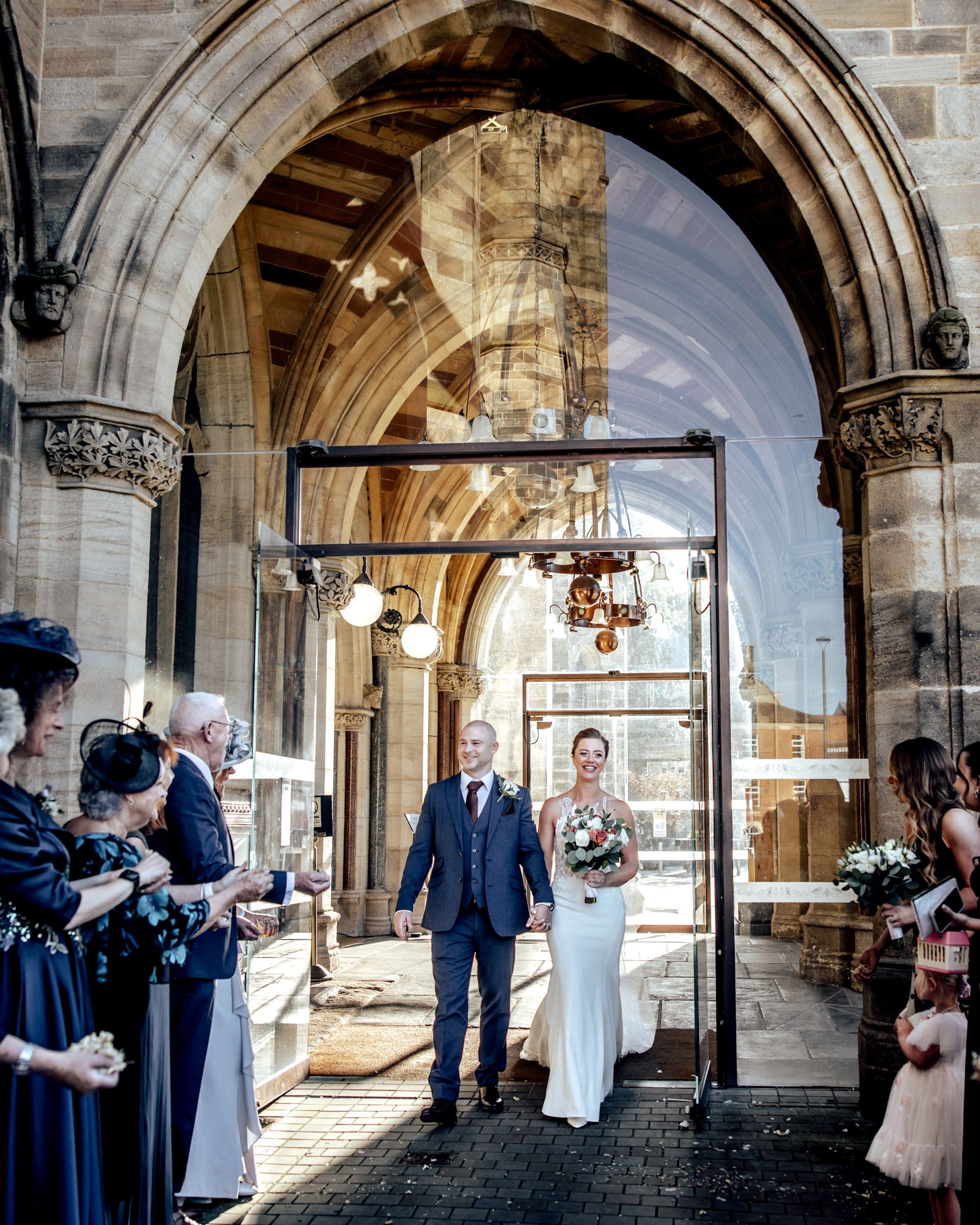 Newlyweds stroll hand in hand beneath a grand Gothic stone archway as formally dressed guests look on.