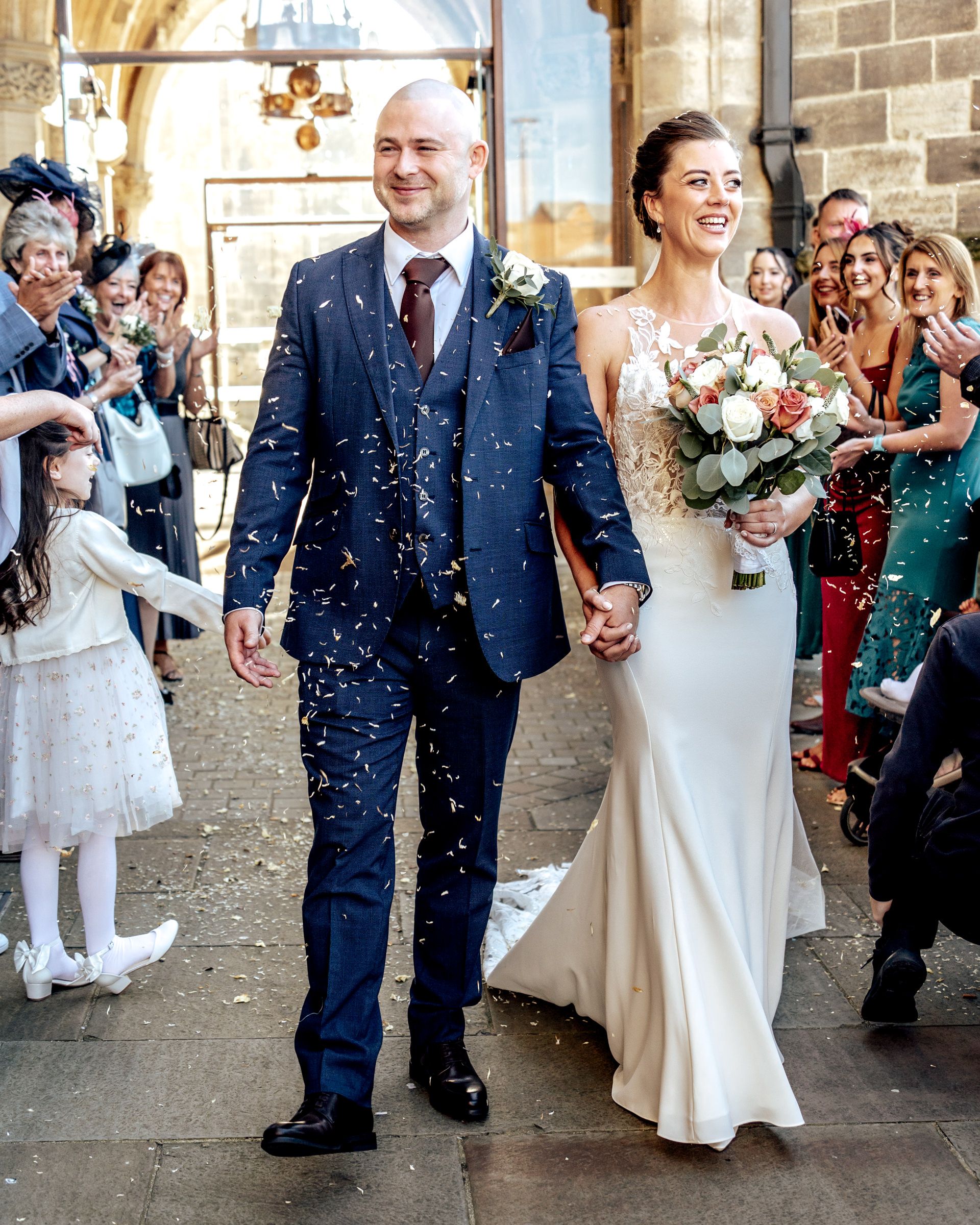 Smiling bride in a white gown and groom in a navy three-piece suit hold hands as wedding guests cheer and throw confetti outside a church.