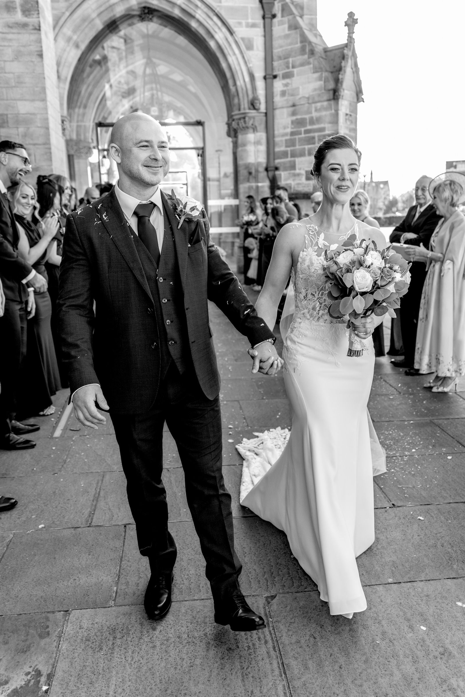 Smiling bride in a fitted lace gown holding a bouquet walks hand in hand with a groom in a dark suit outside a stone church as guests cheer.