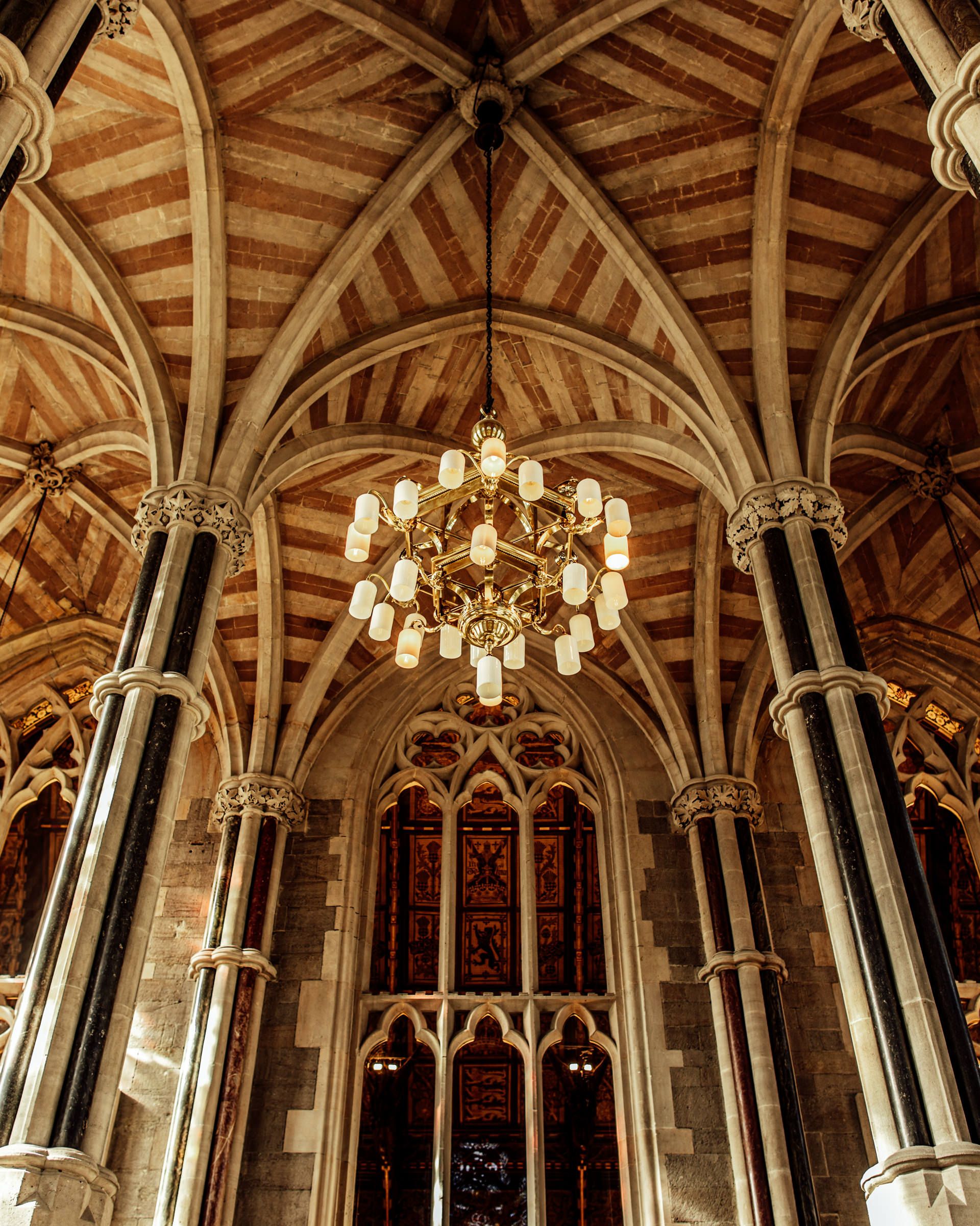 Gothic vaulted ceiling with striped ribbed arches, a hanging globe chandelier, stone columns and a tall stained glass window below.