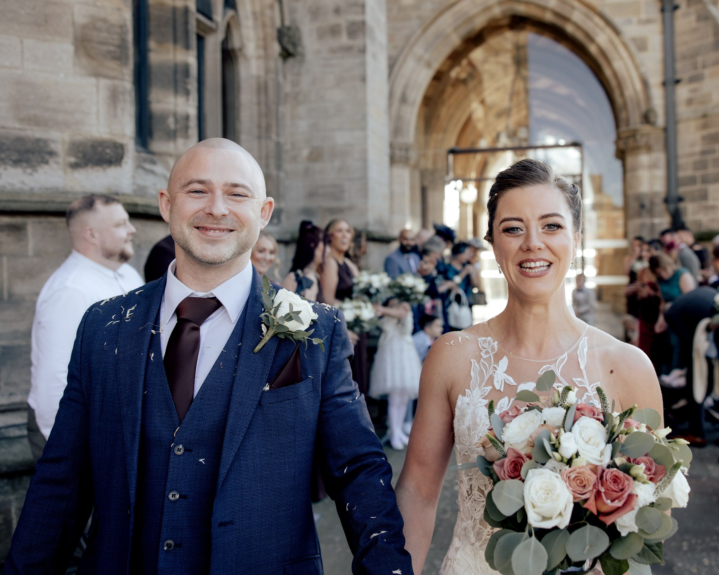 Smiling bride in a lace gown holding a pink and white rose bouquet beside a groom in a navy suit outside a stone church with guests behind.