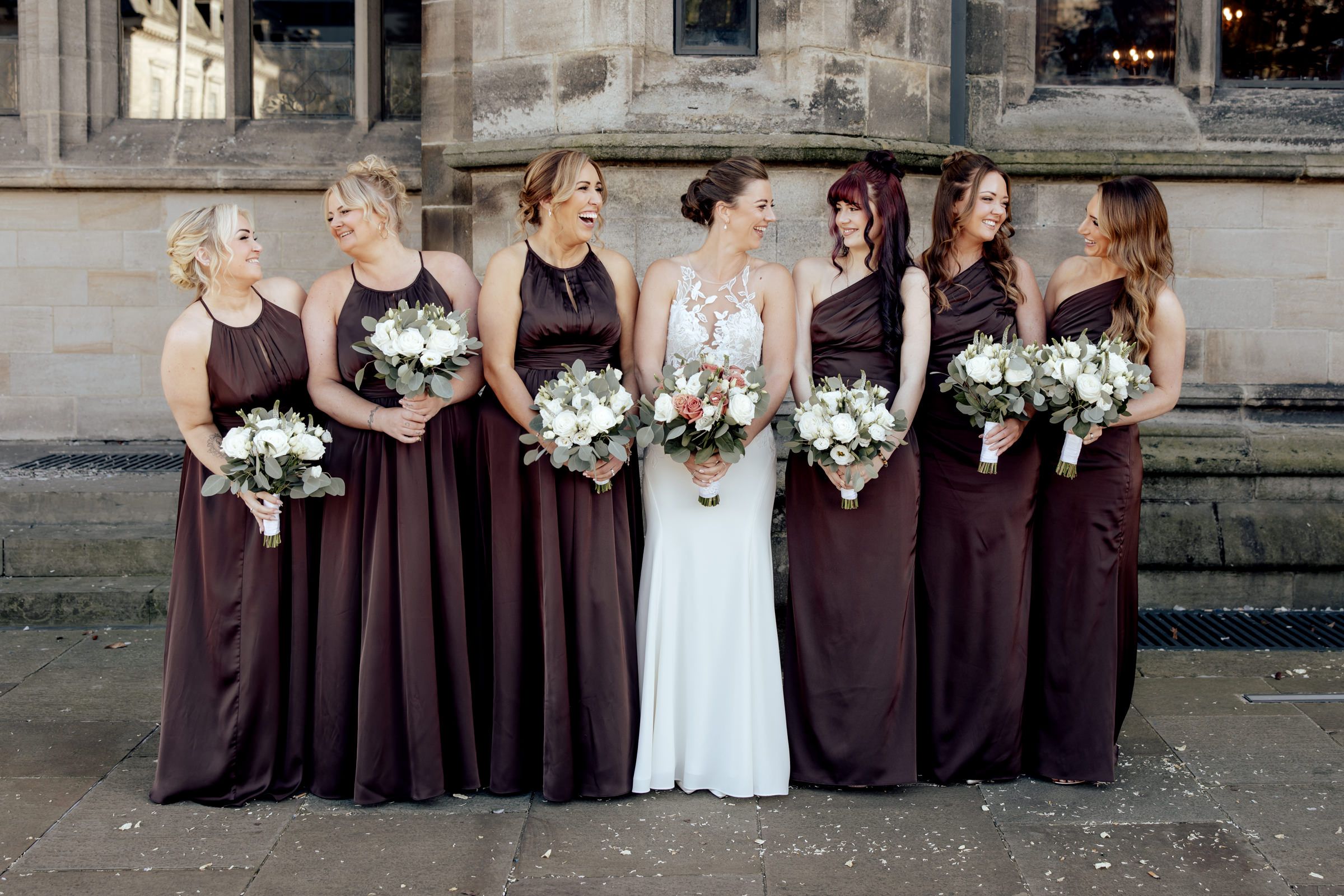 Bride in a white gown laughs with six bridesmaids in long burgundy dresses, all holding white and blush bouquets outside a stone building.