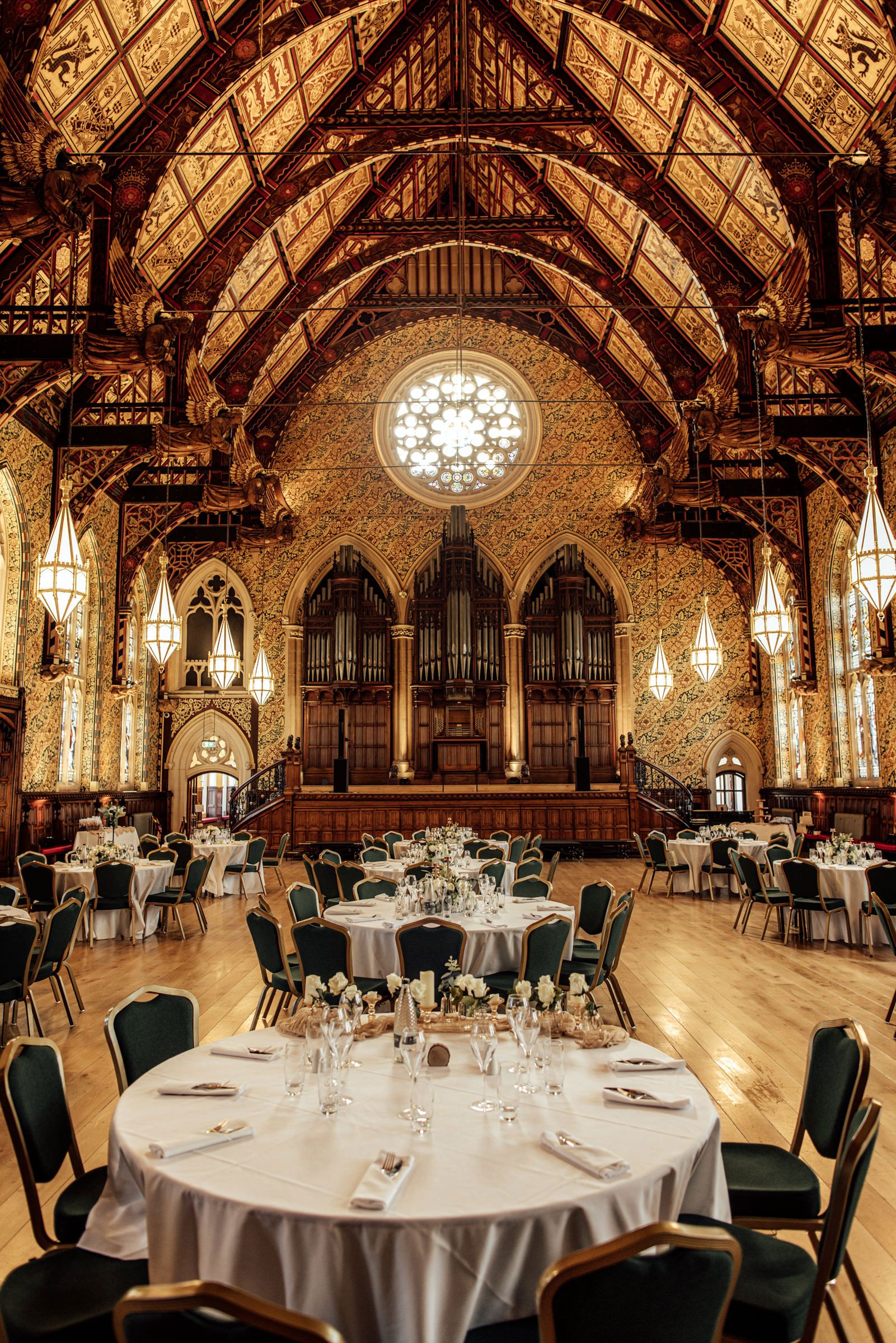 Elegant banquet hall with round tables set for a formal event beneath a vaulted wooden ceiling, pipe organ, and rose window.