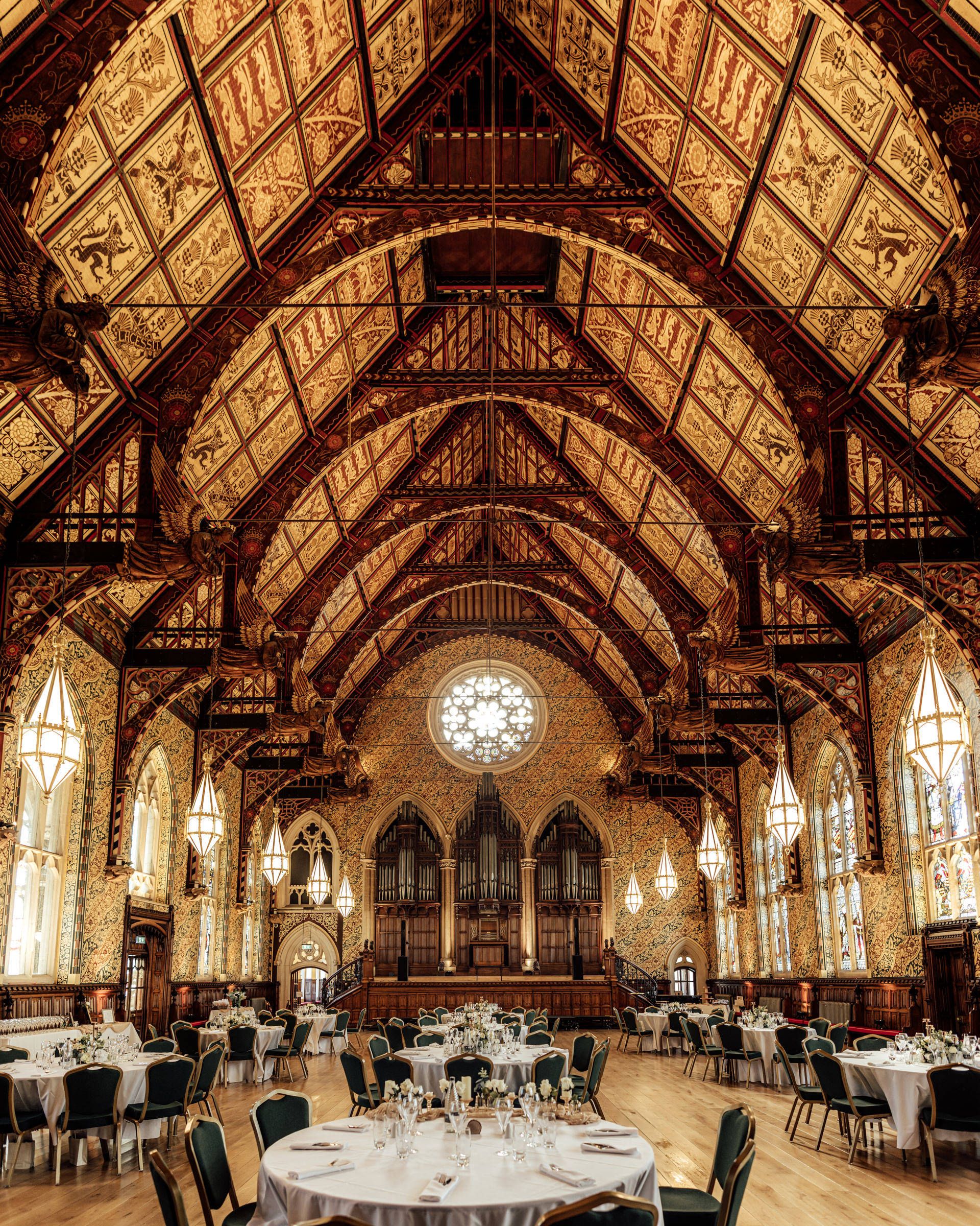 Grand hall with ornate timber hammerbeam roof, rose window and pipe organ, set with round banquet tables for an elegant event.
