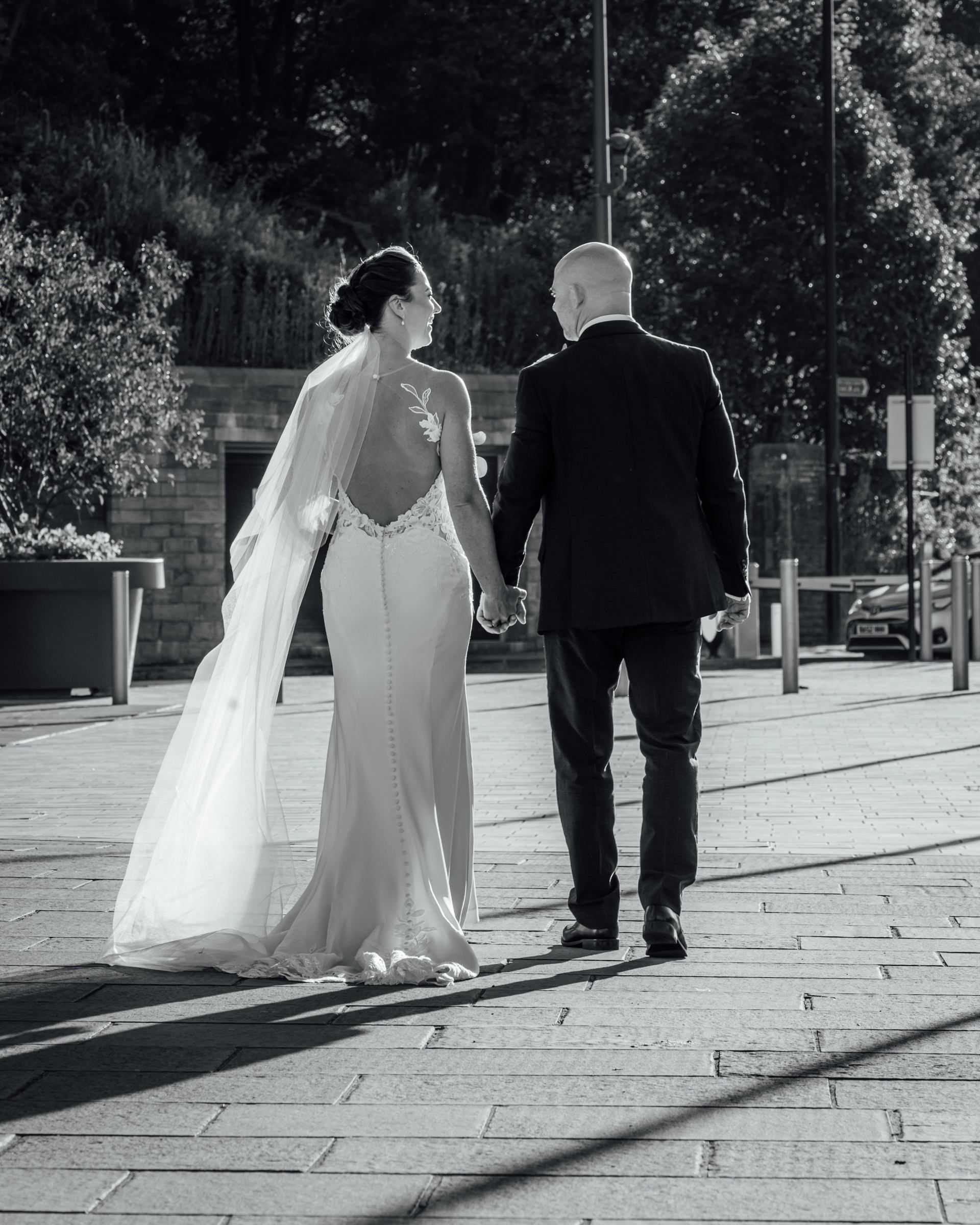 Black and white photo of a bride in a backless gown with a long veil holding hands with her groom as they walk down a sunlit paved street.