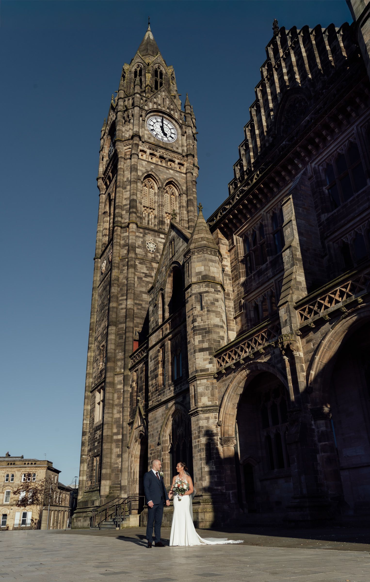 Bride and groom pose on a sunlit plaza before a tall Gothic stone clock tower and historic town hall under a clear blue sky.