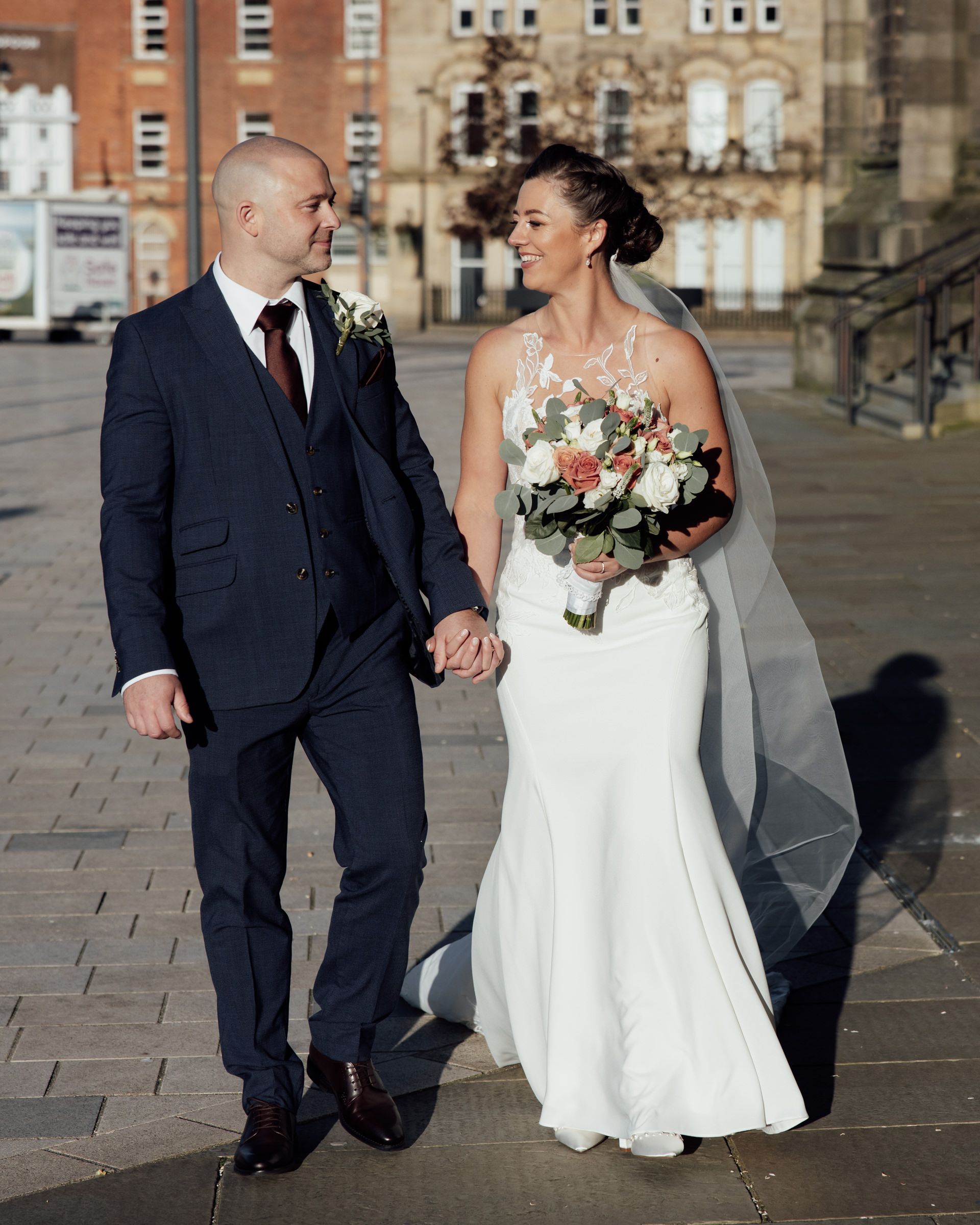 Bride in a white gown with veil and bouquet walks hand in hand with a groom in a navy suit across a sunlit stone plaza.
