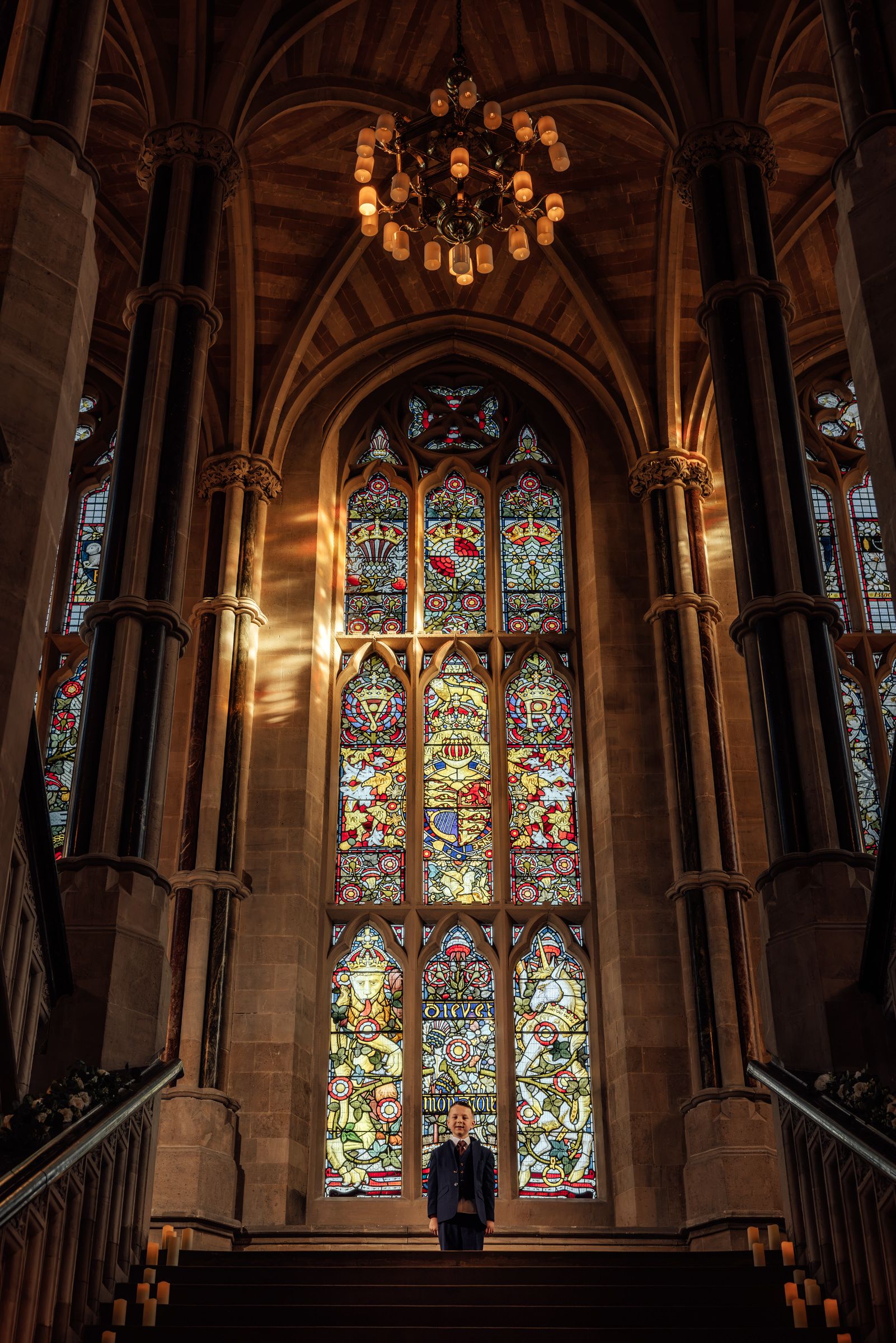 Person stands before a tall Gothic stained glass window in a grand stone hall with a chandelier, sunlight and a staircase.