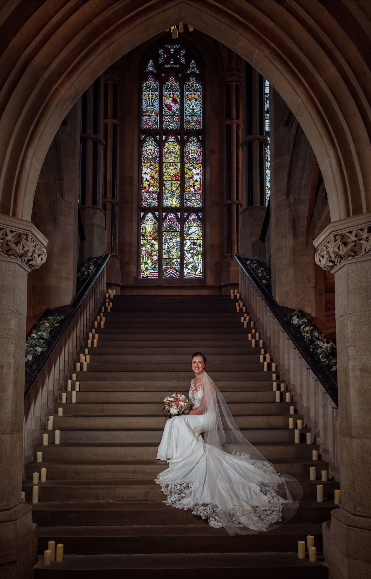 Bride in a flowing white gown and veil sits on a grand stone staircase lined with candles beneath a stunning stained glass window.