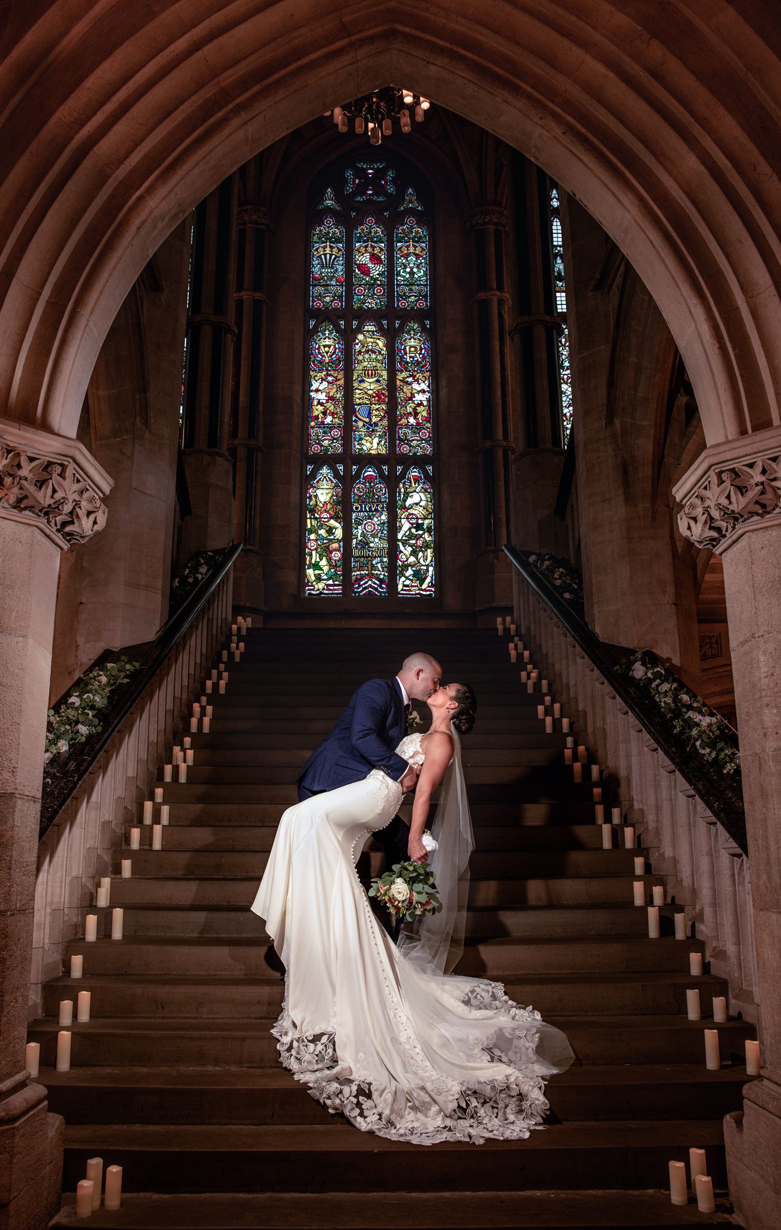 Bride in a lace-trimmed white gown and groom in a navy suit share a dip kiss on candlelit stone stairs beneath a stained glass window.