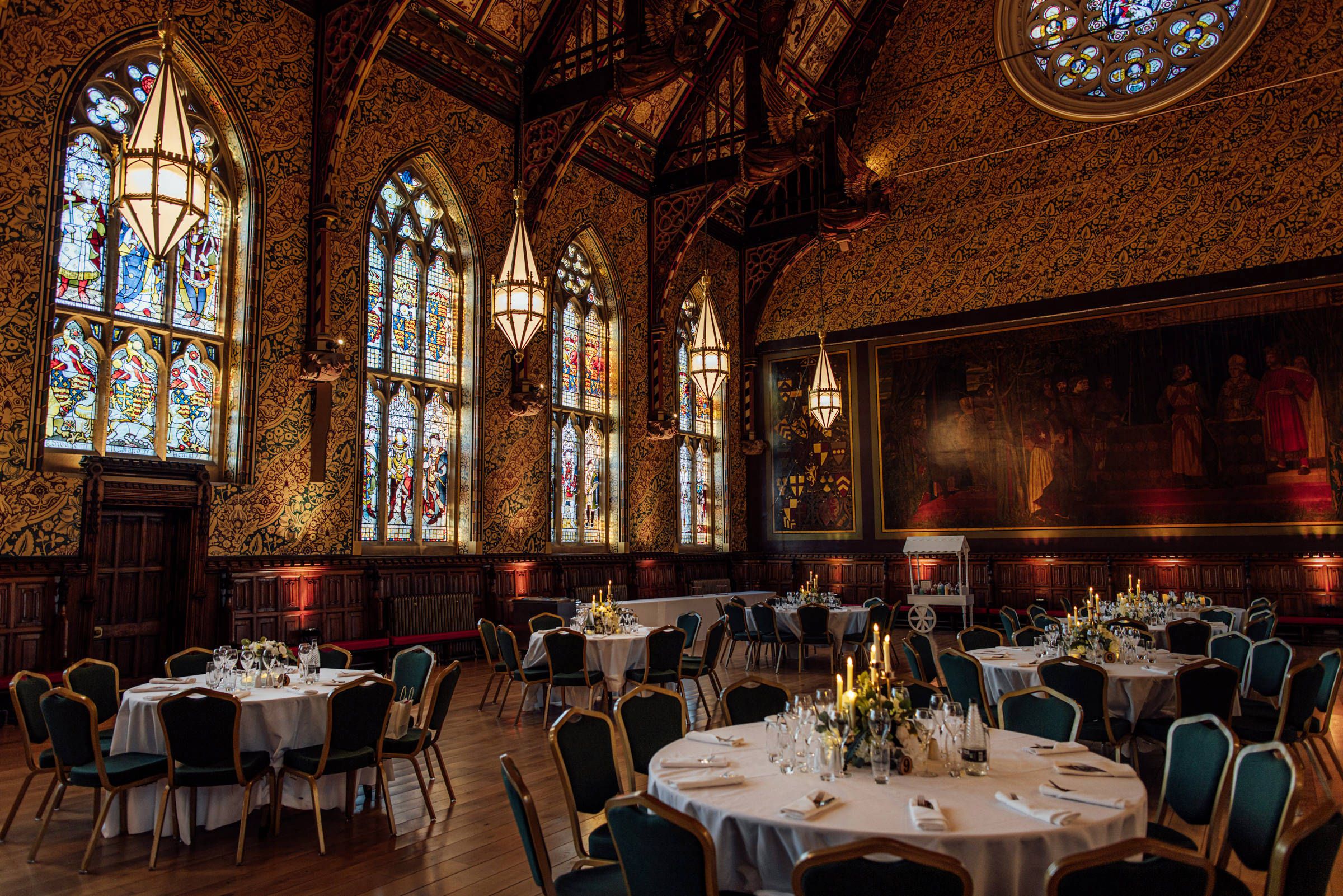 Elegant banquet hall with round tables, teal chairs, stained glass windows, pendant lanterns and ornate wood-paneled walls.