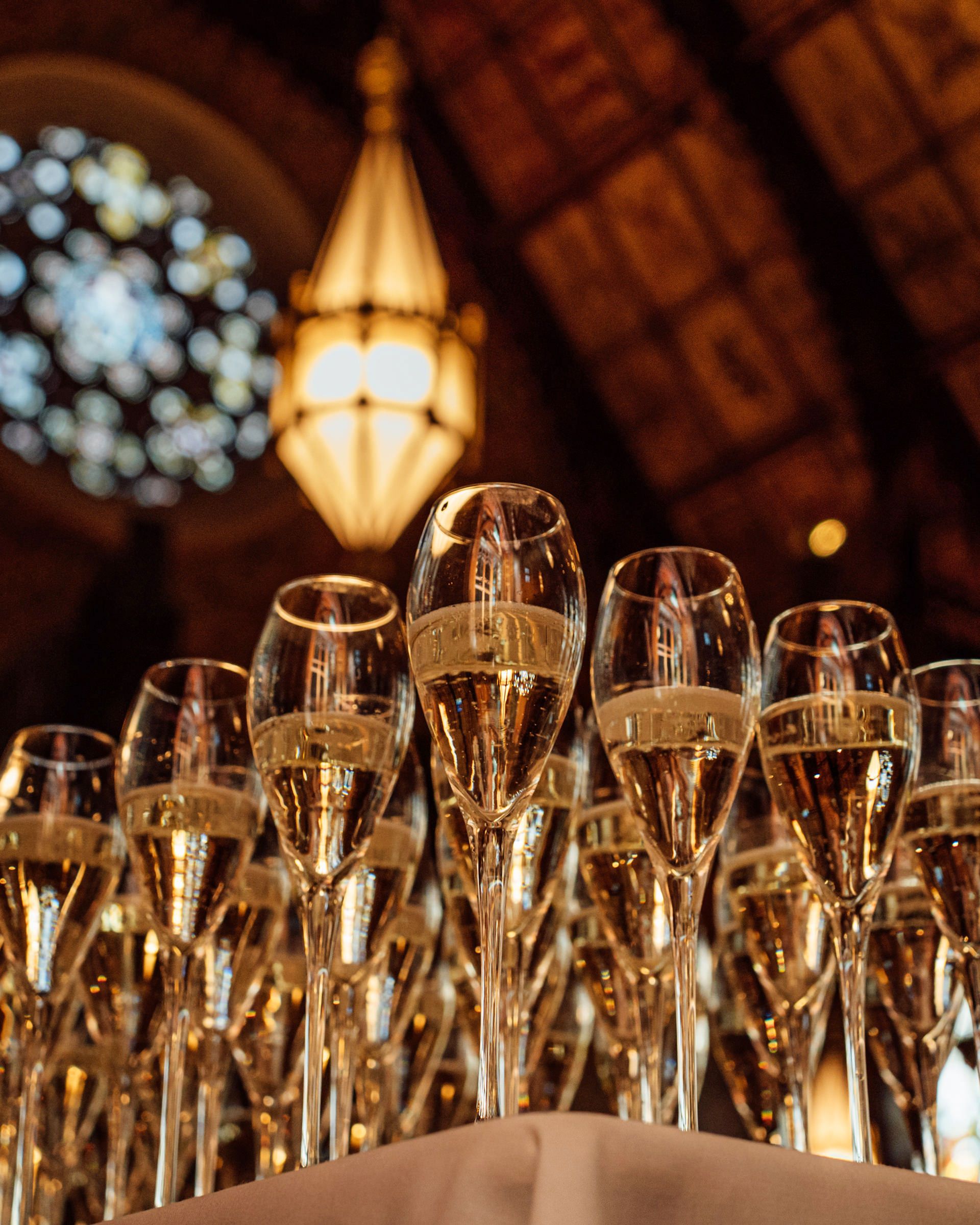Rows of champagne flutes filled with sparkling wine on a table beneath a glowing pendant lamp and ornate stained glass window.