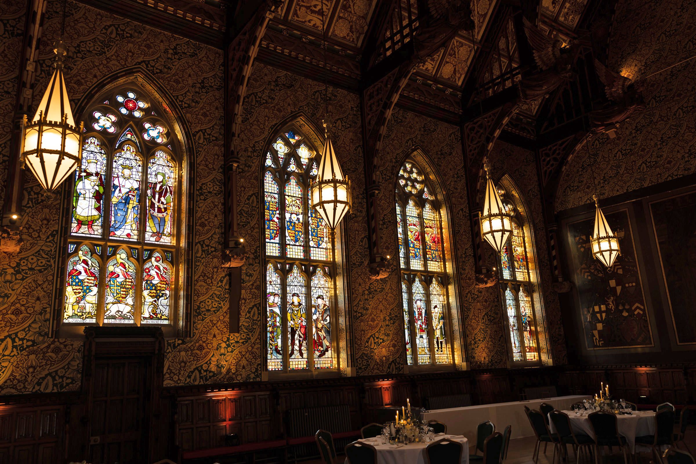 Ornate Gothic hall with glowing stained glass windows, hanging lanterns, carved wooden beams, and elegantly set dining tables.