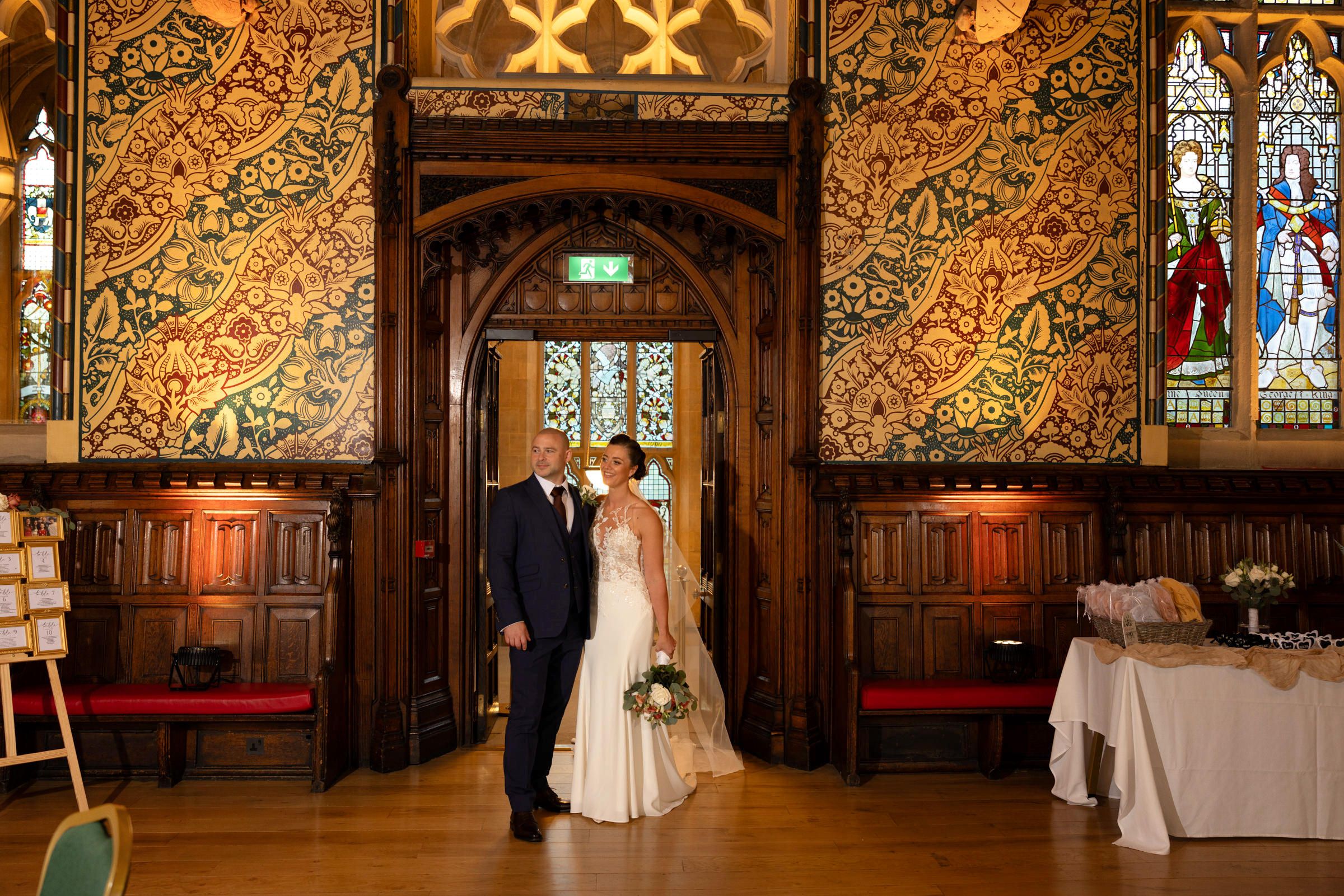Bride in a white gown and groom in a dark suit pose together in an ornate wedding venue with stained glass windows and carved wood paneling.