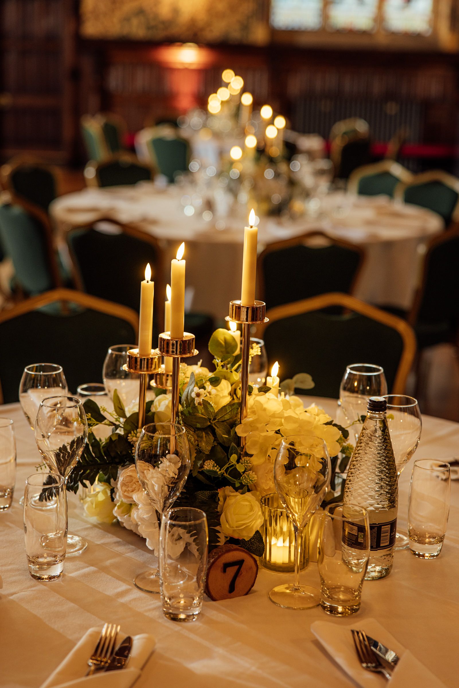Elegant wedding reception table with lit taper candles, yellow rose centerpiece, glassware, and table number in a warmly lit banquet hall.