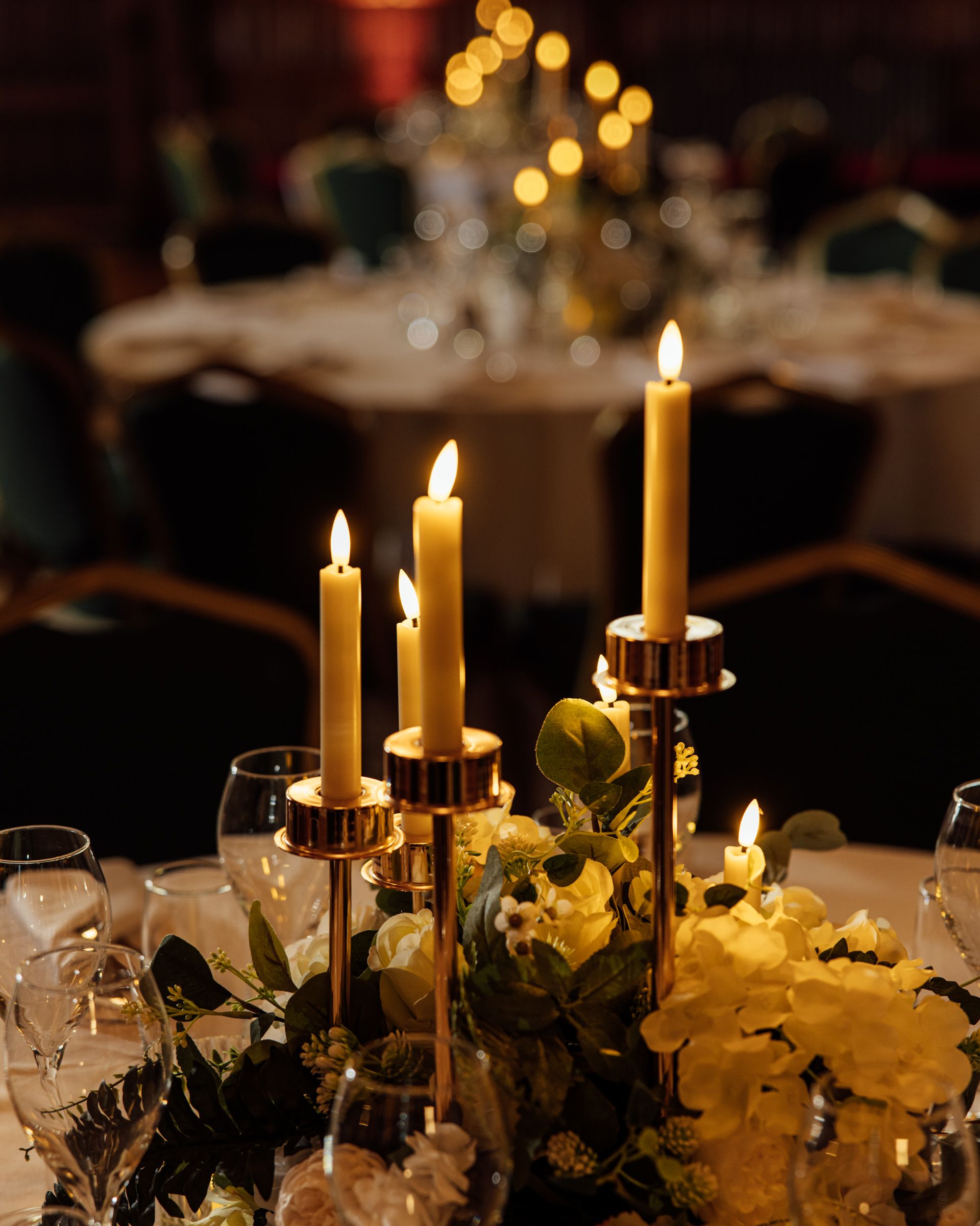 Lit ivory taper candles in gold candle holders with white roses and greenery centerpiece on an elegant banquet table at a wedding reception.