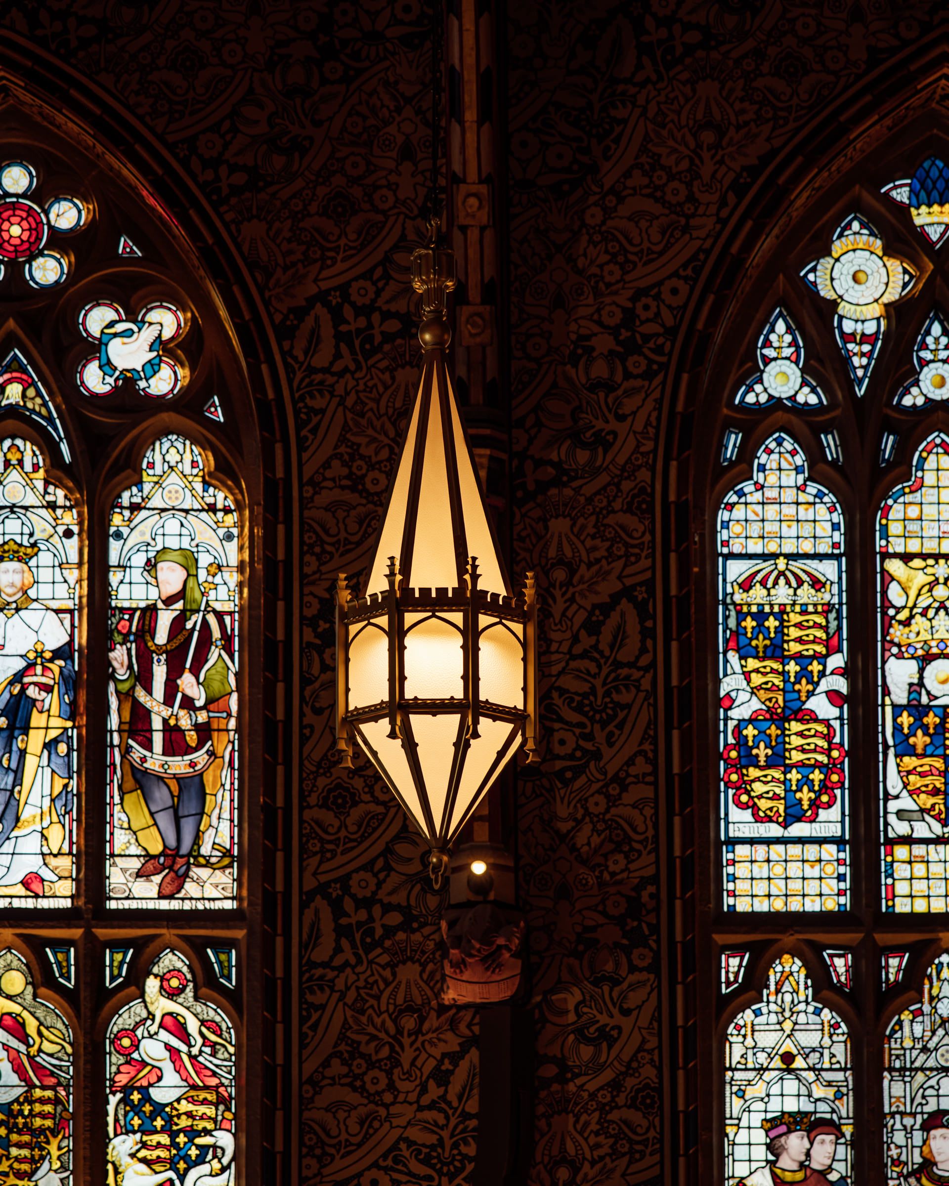 Ornate hanging lantern between two Gothic stained glass windows with heraldic shields and robed figures against carved dark wood walls.